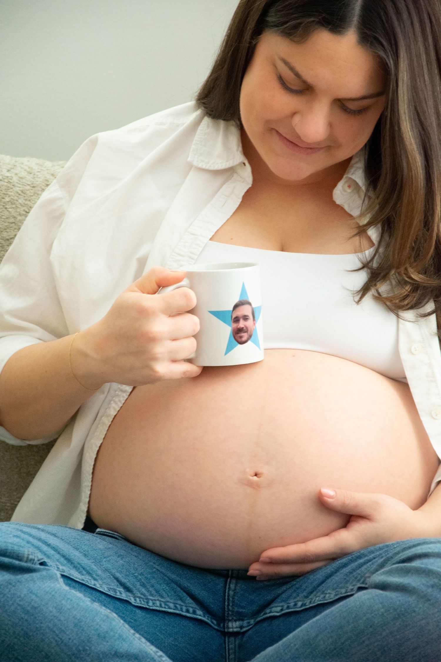 Expectant mother in white shirt and jeans holds coffee mug while gently touching her pregnant belly. Professional maternity photography