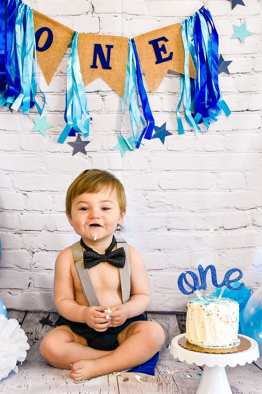 Small boy wearing a bow tie and suspenders smiling under a blue banner that reads 