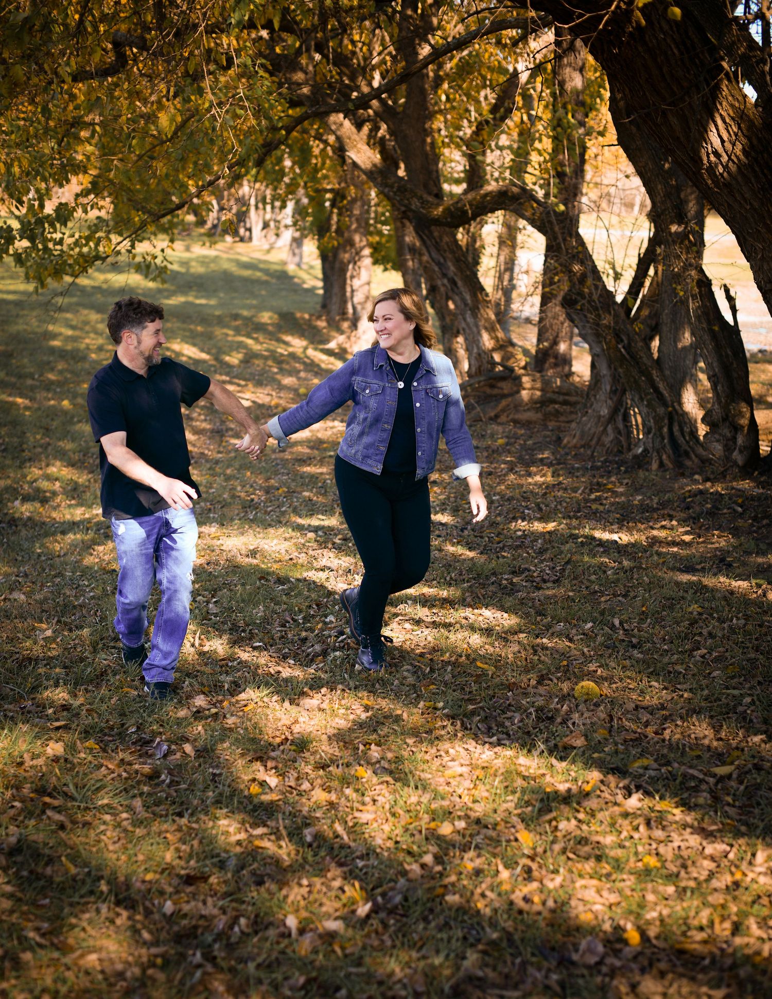 Couple holding hands and walking playfully under large tree in sunlit autumn park setting.
