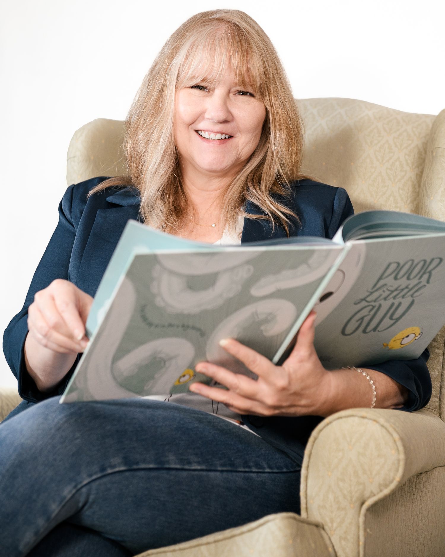 Smiling woman reading Poor Little Guy children's book while seated in a cozy armchair.