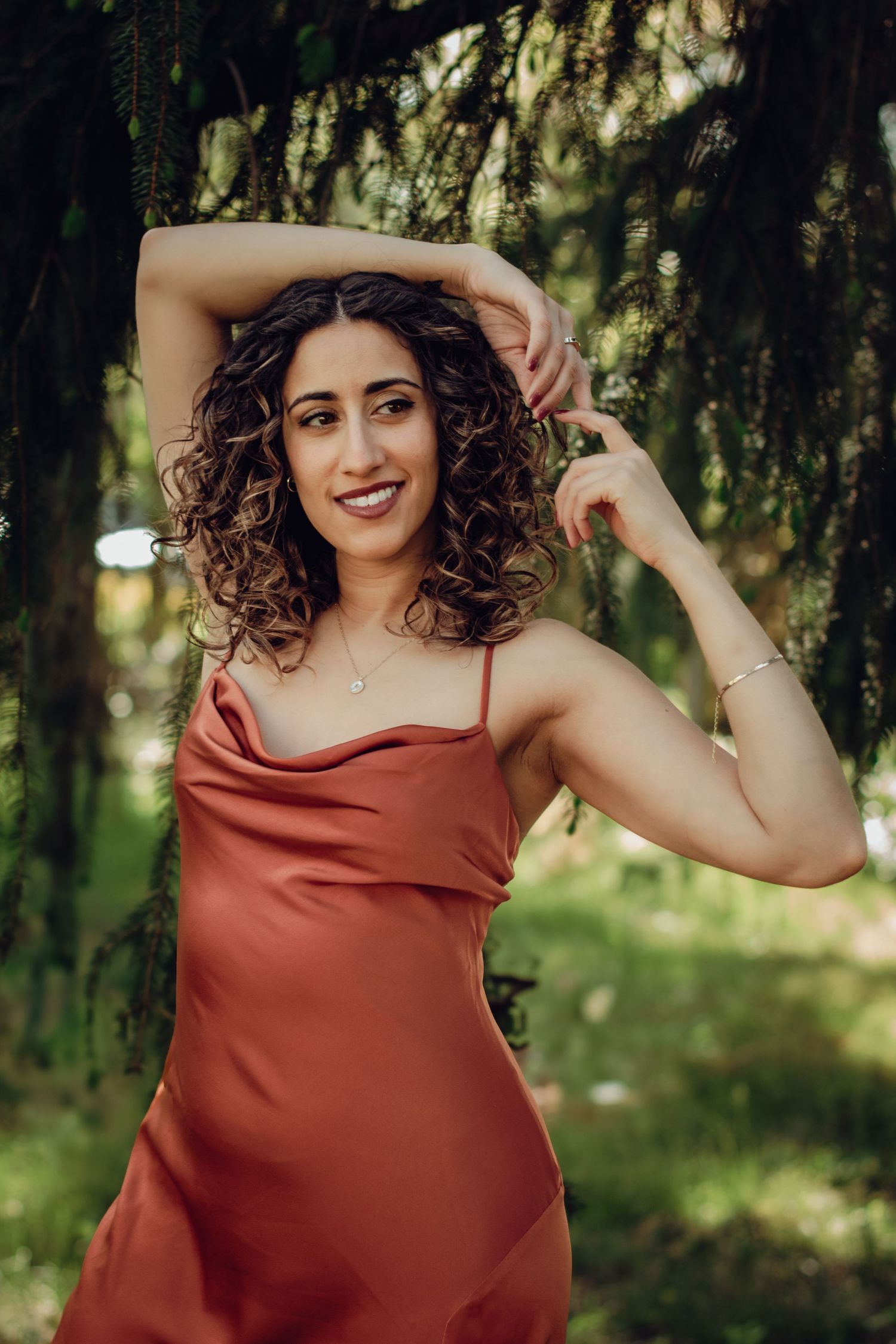 Woman in orange dress poses outdoors with natural curly hair, surrounded by greenery and sunlit trees. Professional portrait photography