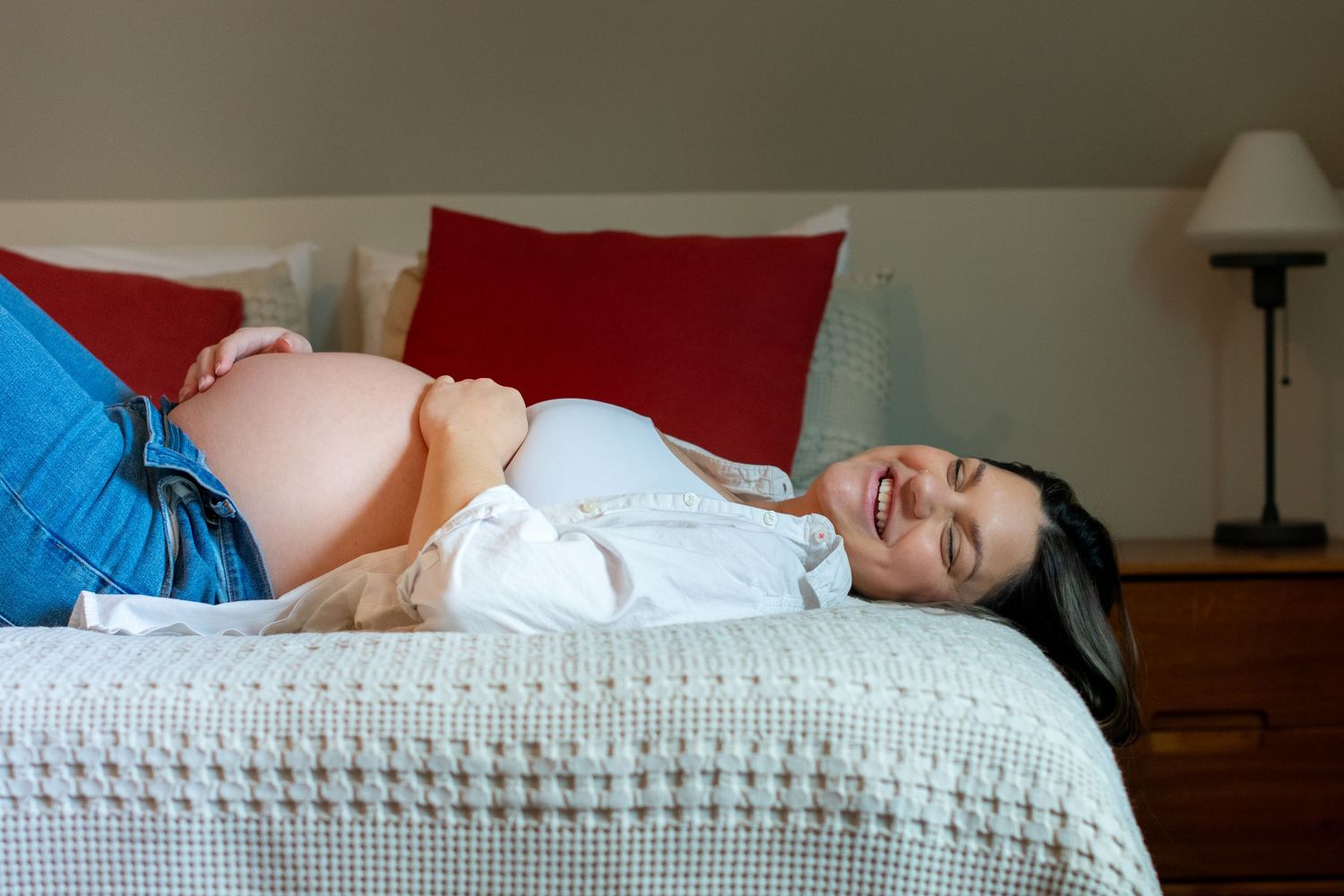 Maternity photo showing woman laughing on white bedding and holding belly in a bedroom setting.