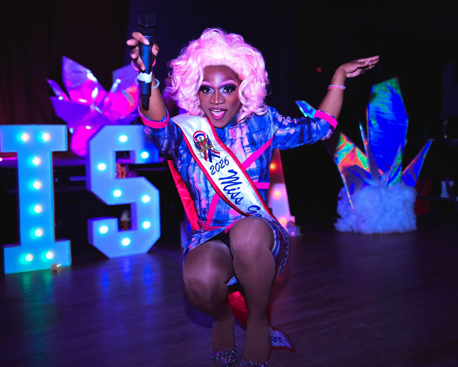 Drag performer in pink wig and sequined outfit posing energetically on stage at colorful nightclub event.