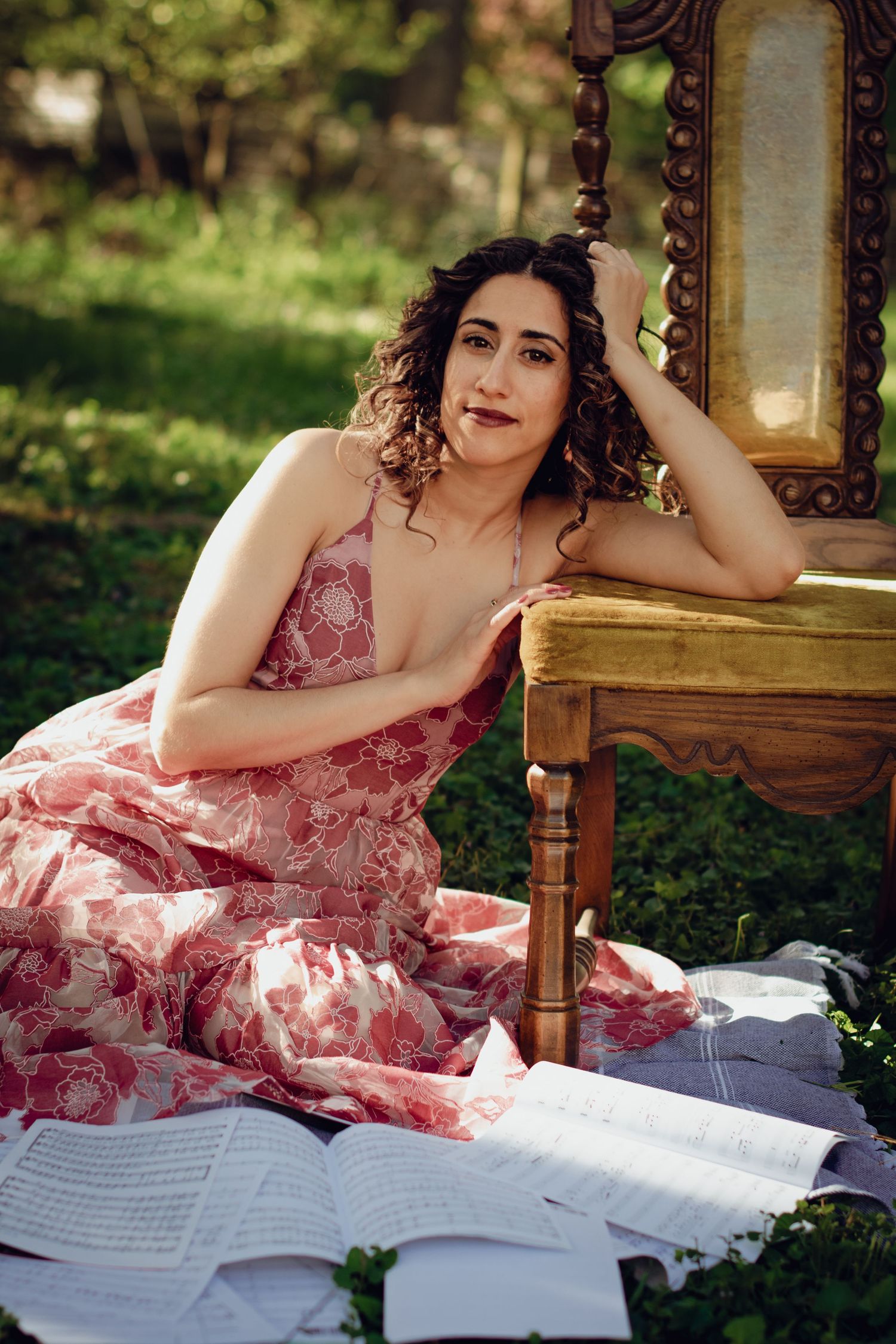 A woman in a floral pink dress lounges on a chair in grass surrounded by sheet music in a sunny garden setting. Professional portrait.