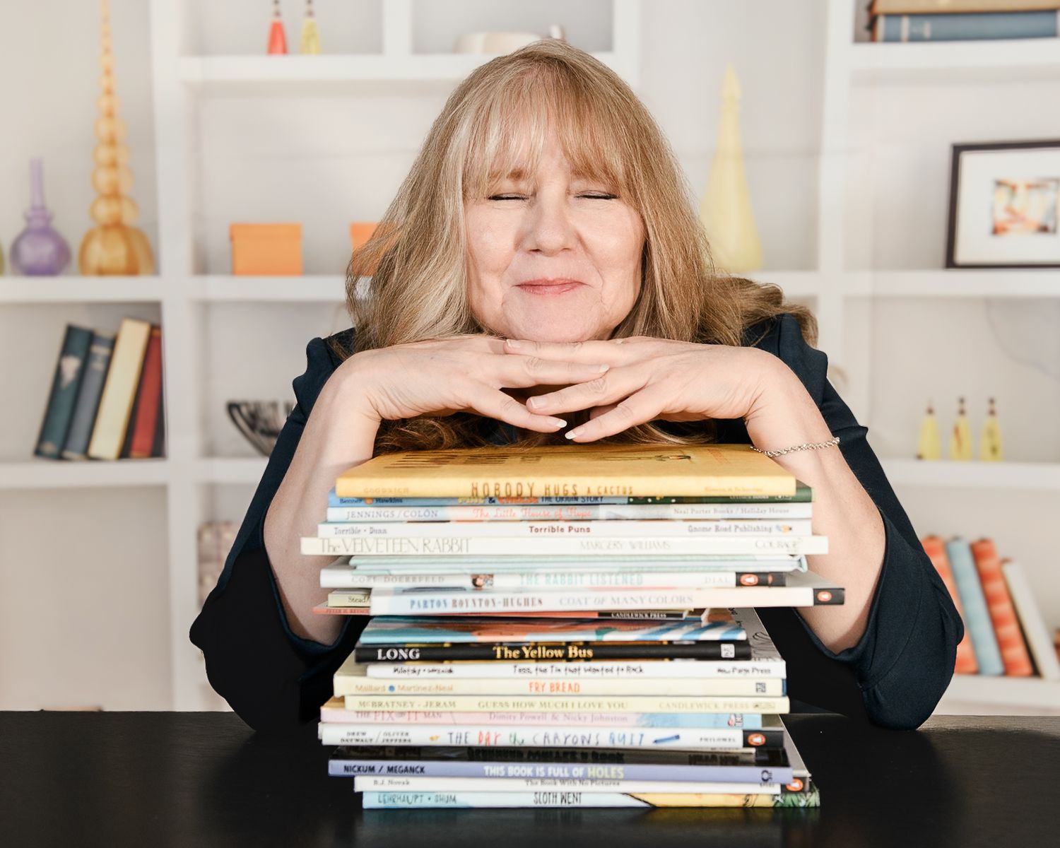 Blonde woman resting chin on stack of colorful children's books at a desk.