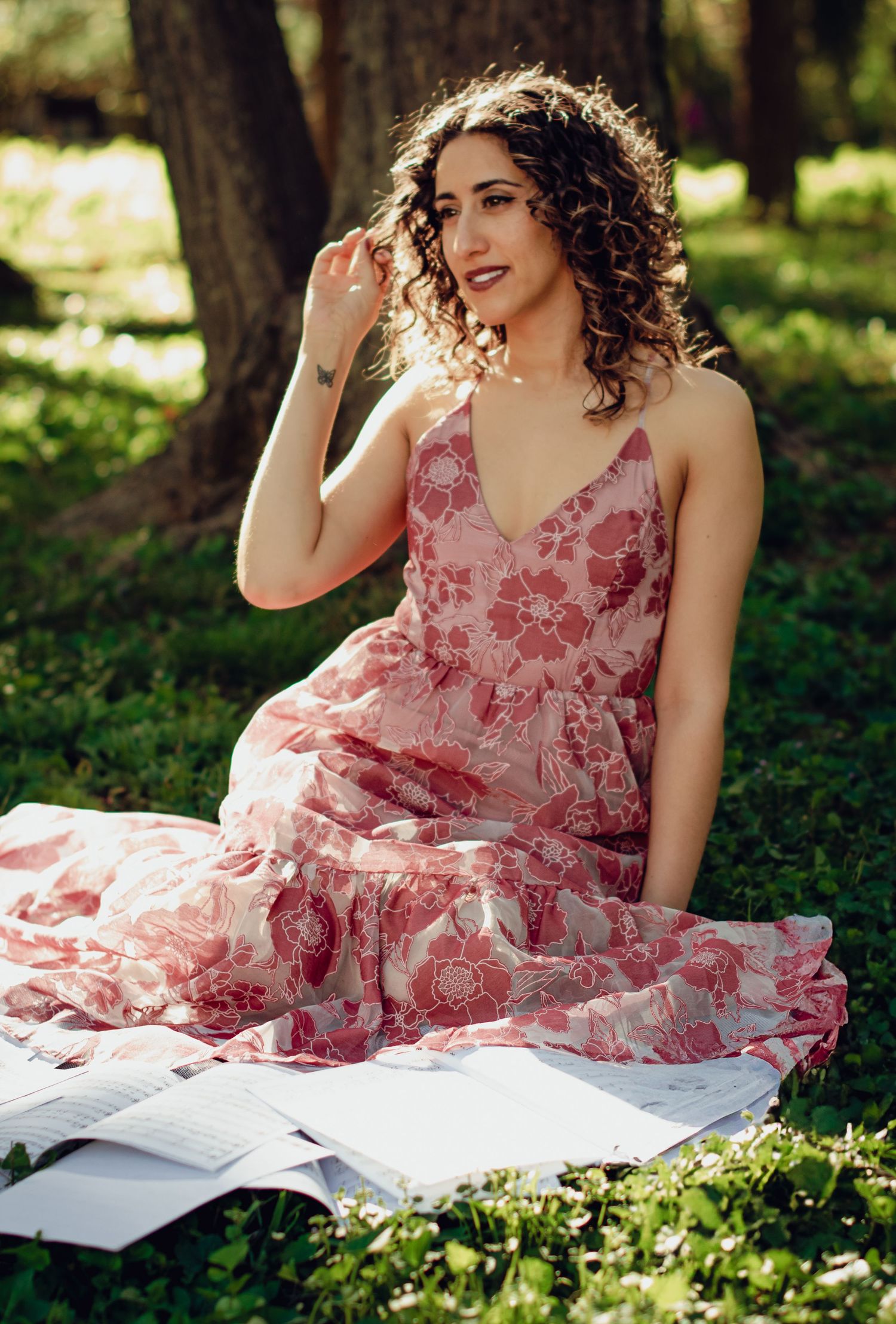 A woman in a floral pink dress sits in grass surrounded by sheet music in a sunny garden setting. Professional portrait photography.