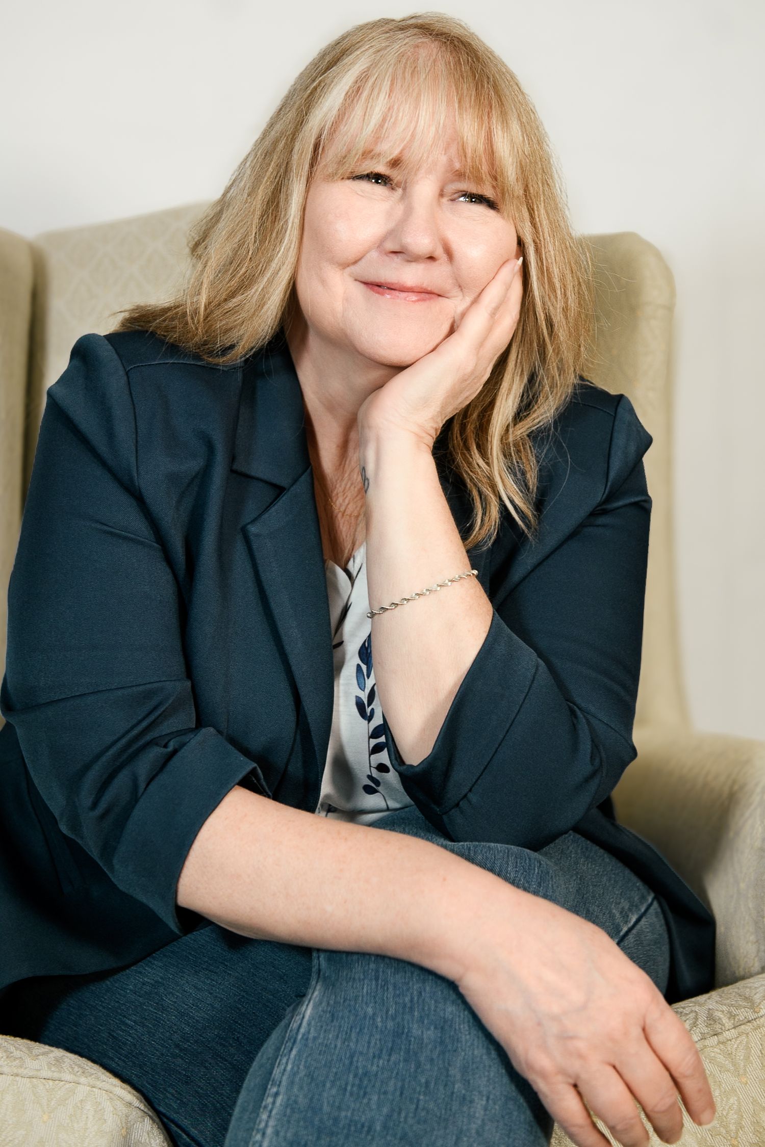 Blonde woman in navy blazer sitting relaxed in armchair with hand on cheek.