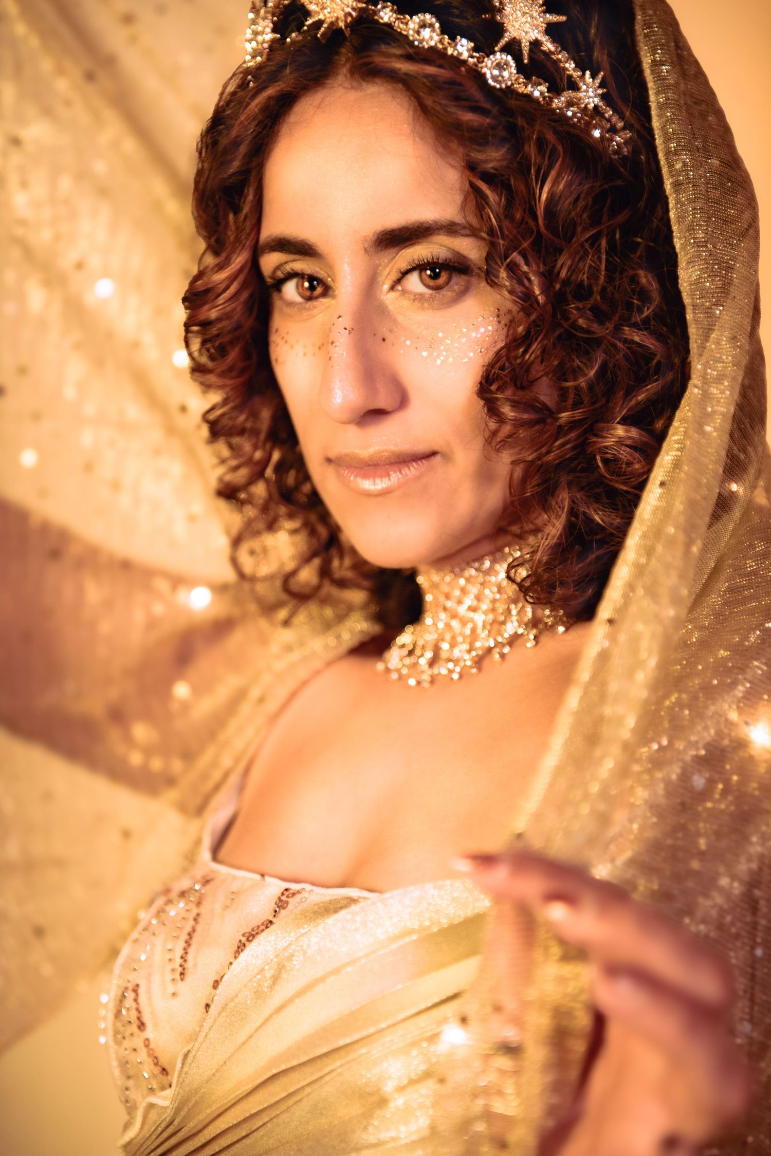 Woman with curly hair wearing gold crown, glitter freckles, and shimmering gold veil in fantasy portrait.