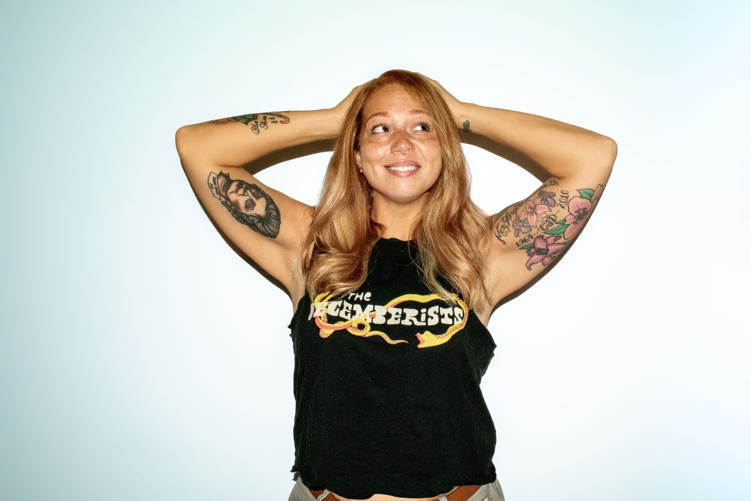 A woman in a sleeveless black graphic tee poses with her hands on her head against a white background. Professional portrait photography