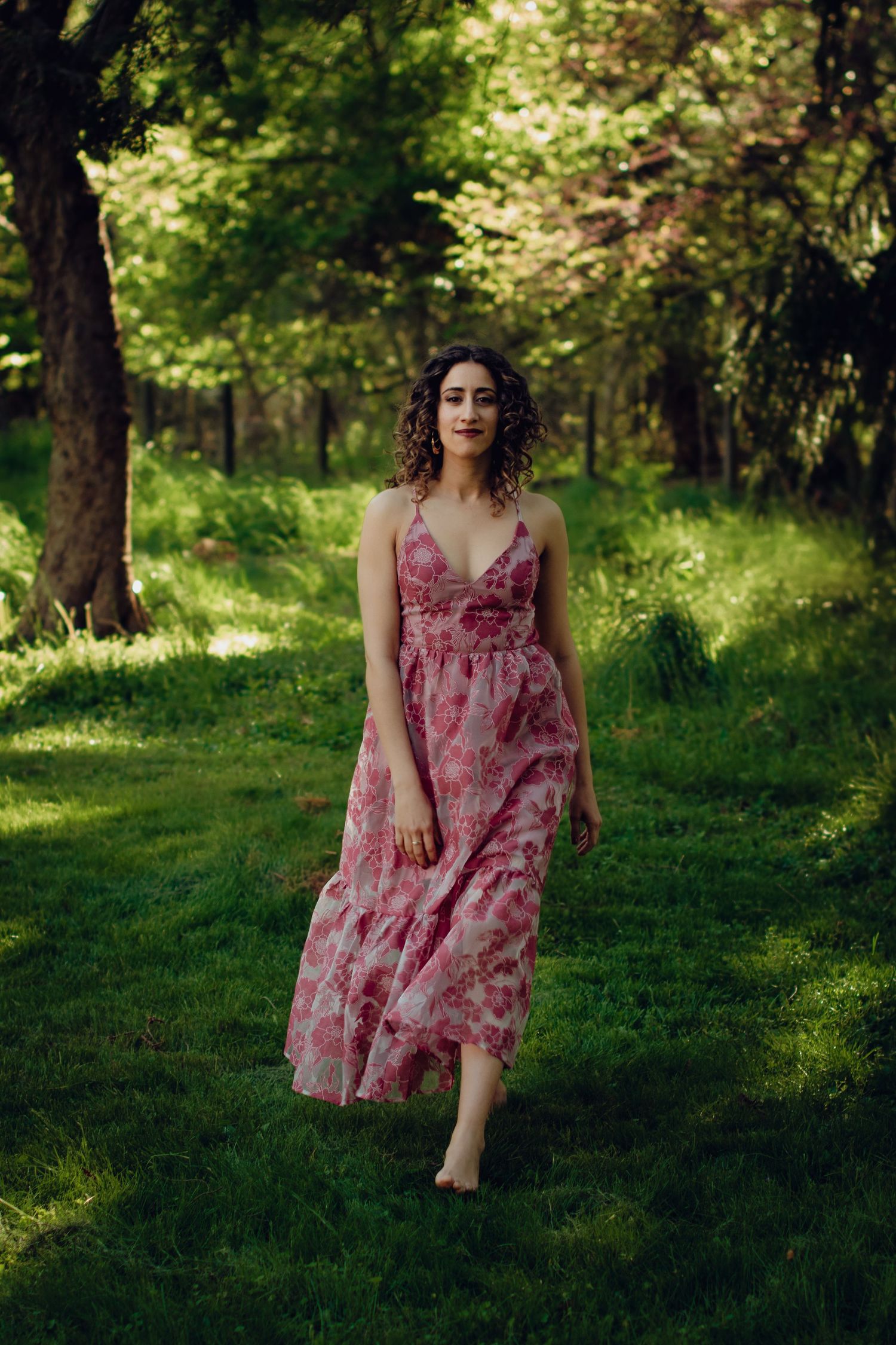 A woman in a flowing pink floral dress walks in a sunlit forest setting with lush green grass and trees. Professional portrait photography