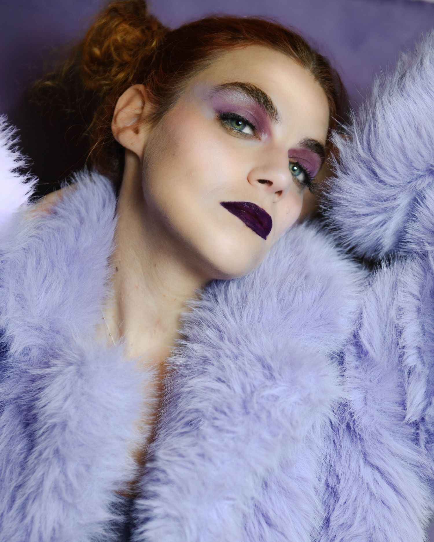 Woman in lavender faux fur coat with bold purple lipstick and dramatic colorful eye makeup posing artistically.