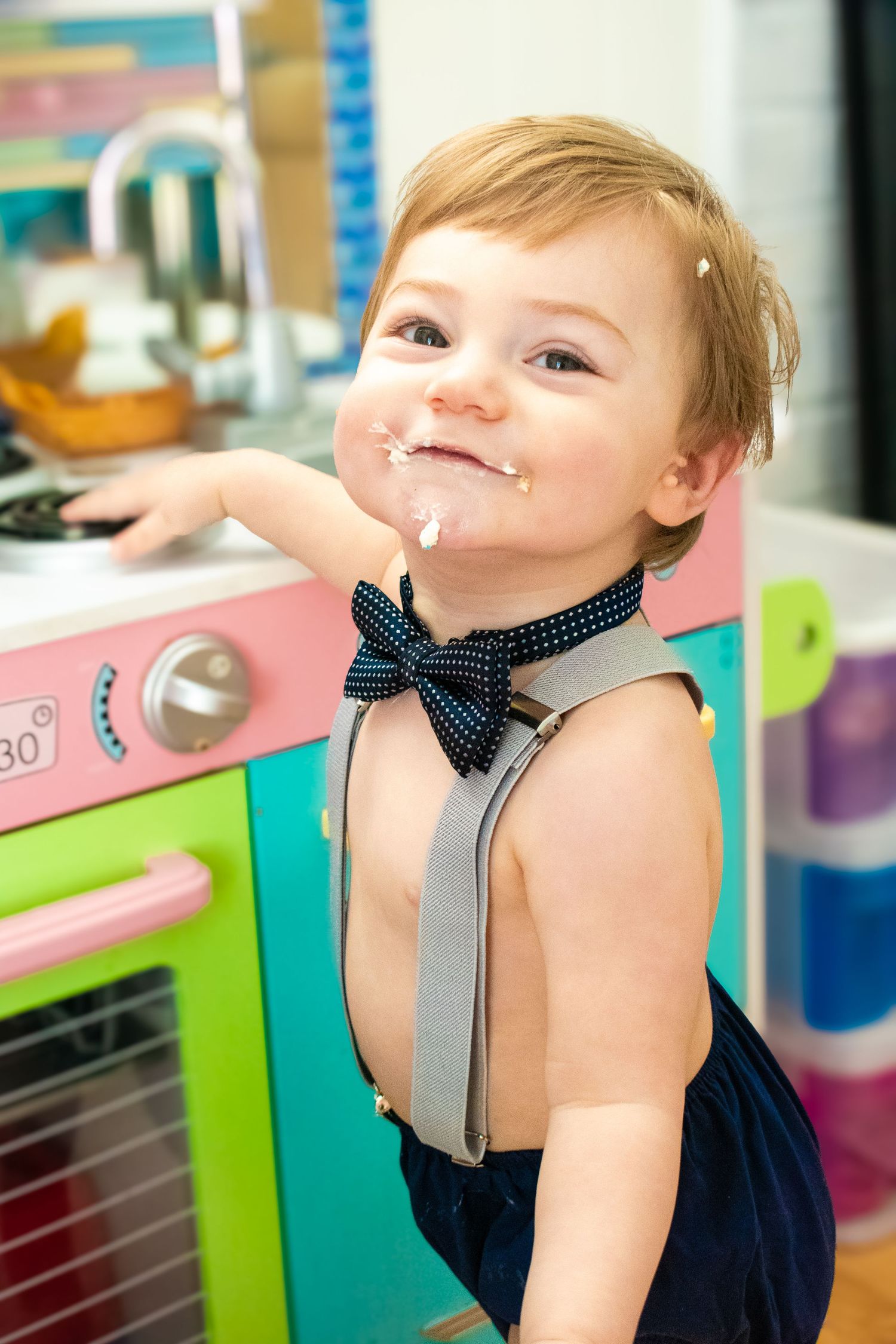 Small boy wearing a bow tie and suspenders smiling with cake on his face. Professional child photography.