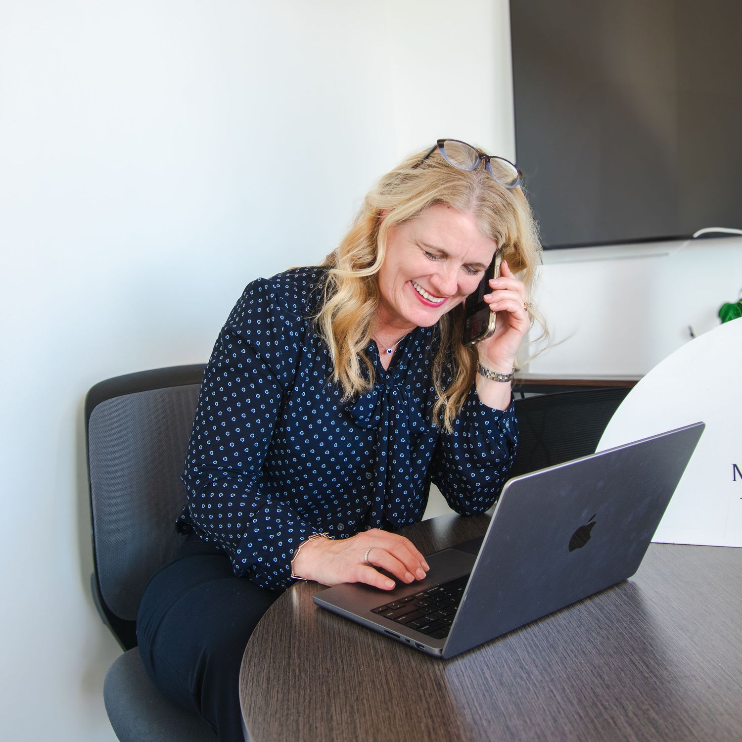 Person working remotely on laptop while seated at desk smiling during phone call.