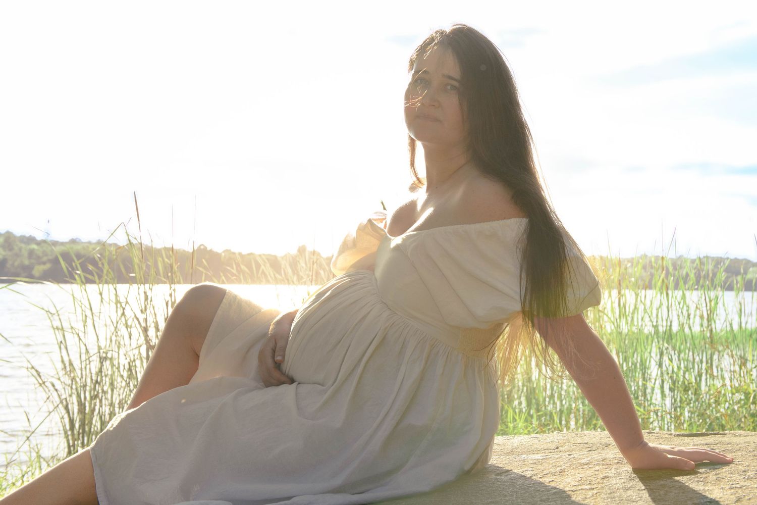 Pregnant woman in off-shoulder white dress sitting outdoors by a lake in bright sunlight.