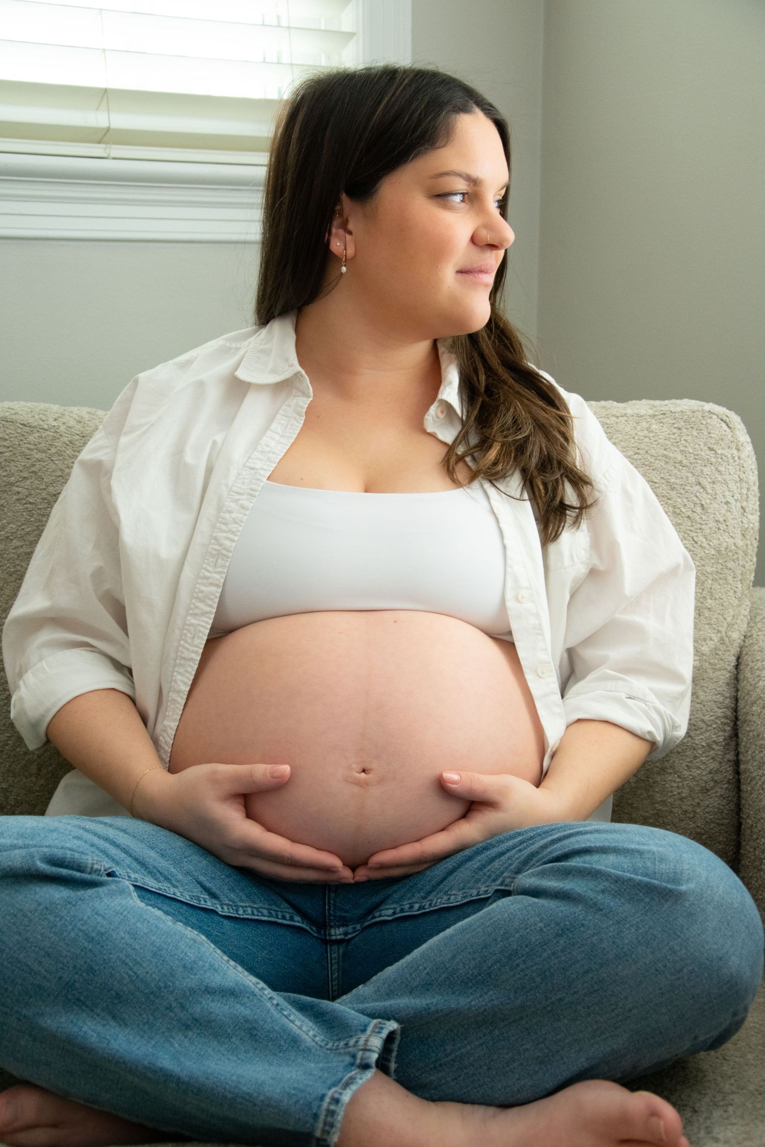 A pregnant woman in a white shirt and jeans sits cross-legged on a couch, gently cradling her belly. Professional maternity photography