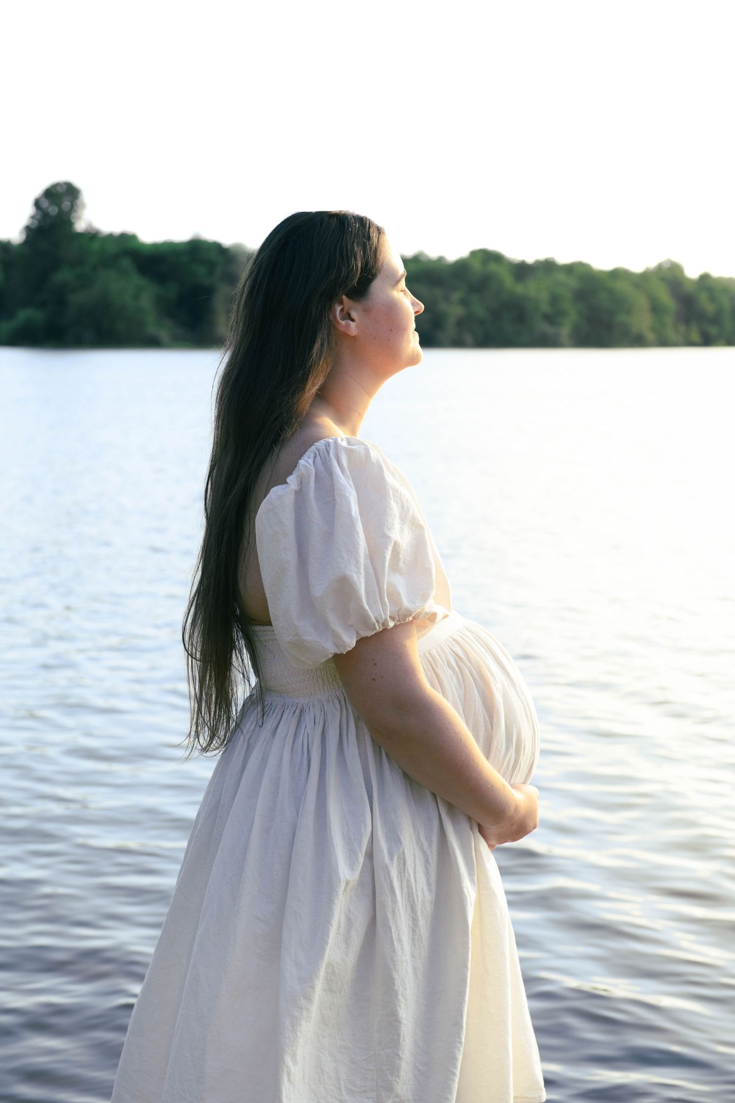 Pregnant woman in white dress standing by a calm lake at sunset, cradling her baby bump.