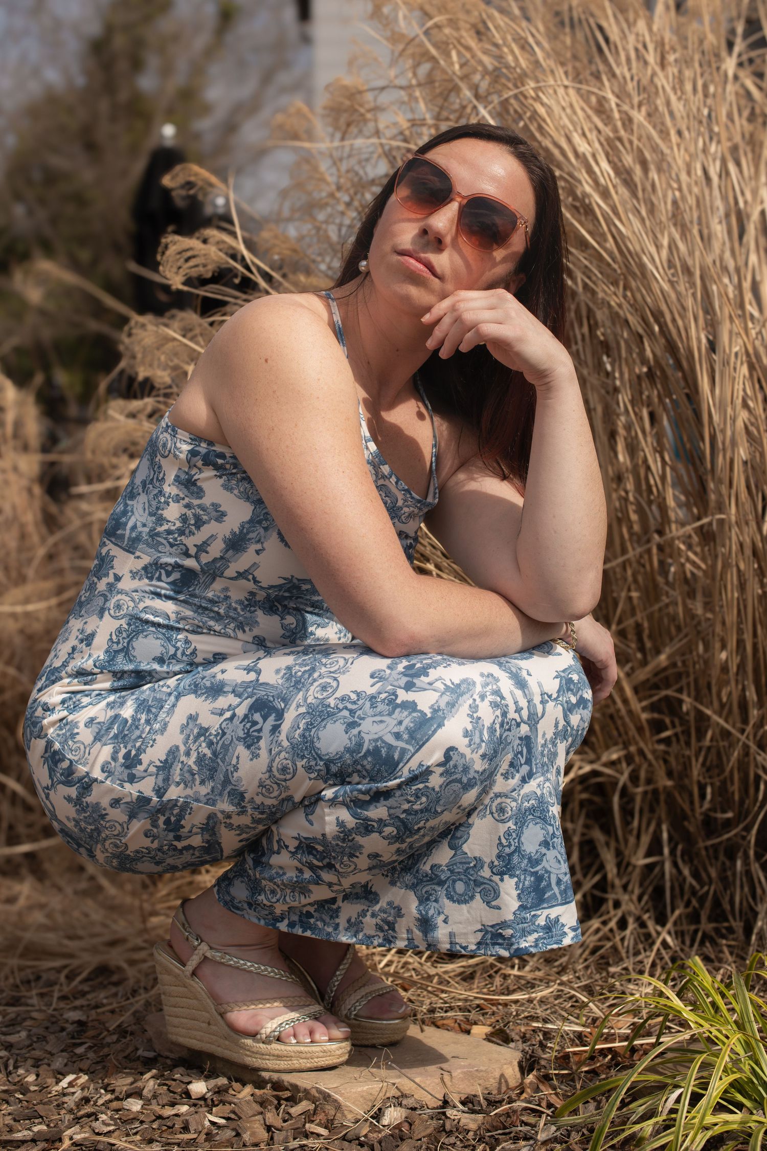 Woman in blue floral jumpsuit and sunglasses crouching outdoors among tall dry grasses on a sunny day.