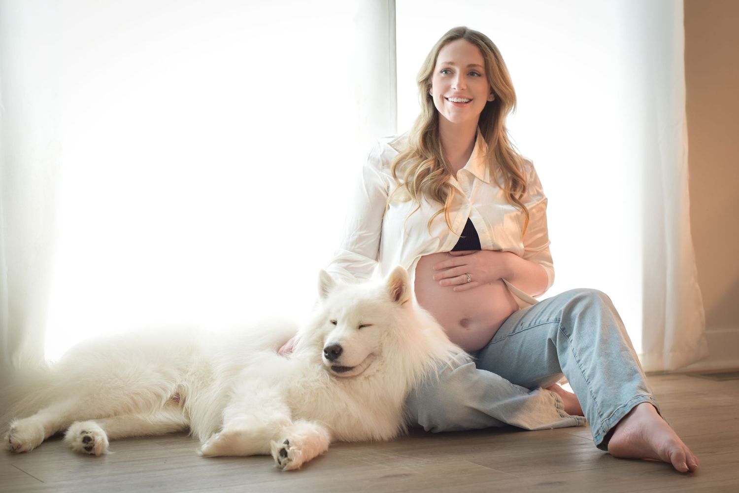 Pregnant woman sitting on floor smiling with a fluffy white Samoyed dog beside her near a bright window.