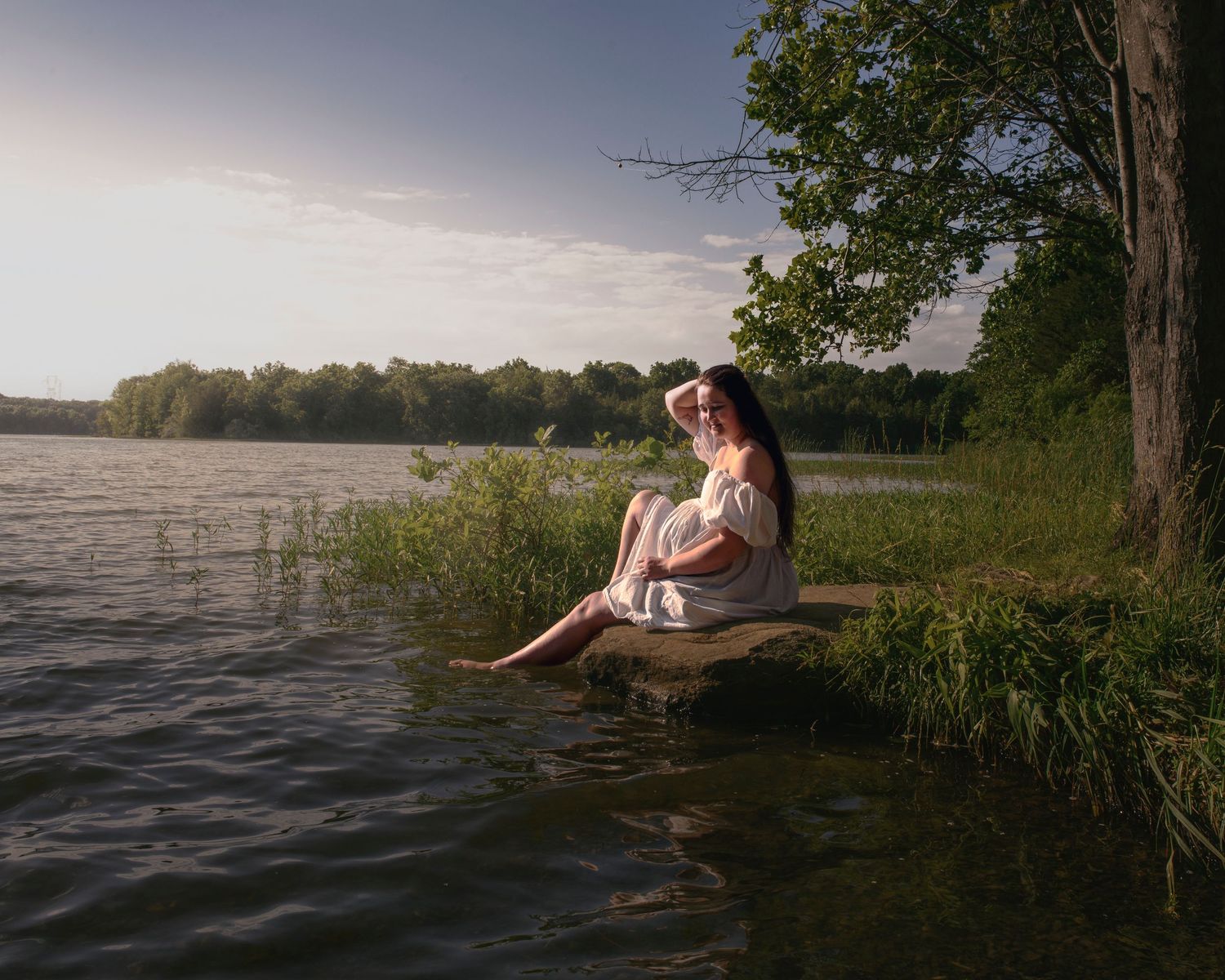 Woman in white dress sitting peacefully on a rock by a tranquil lake at golden hour near lush trees.
