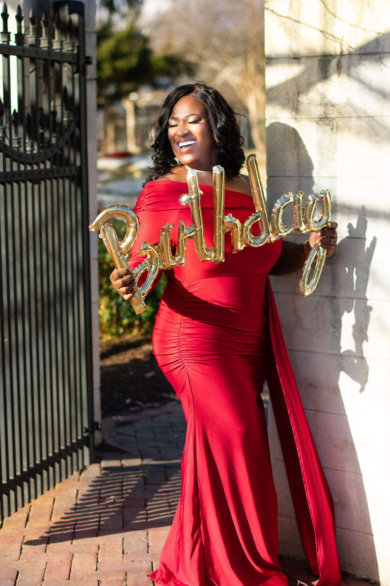 Woman in red off-shoulder gown holding gold 'Birthday' balloon sign. Elegant celebration portrait with dramatic afternoon lighting and archi
