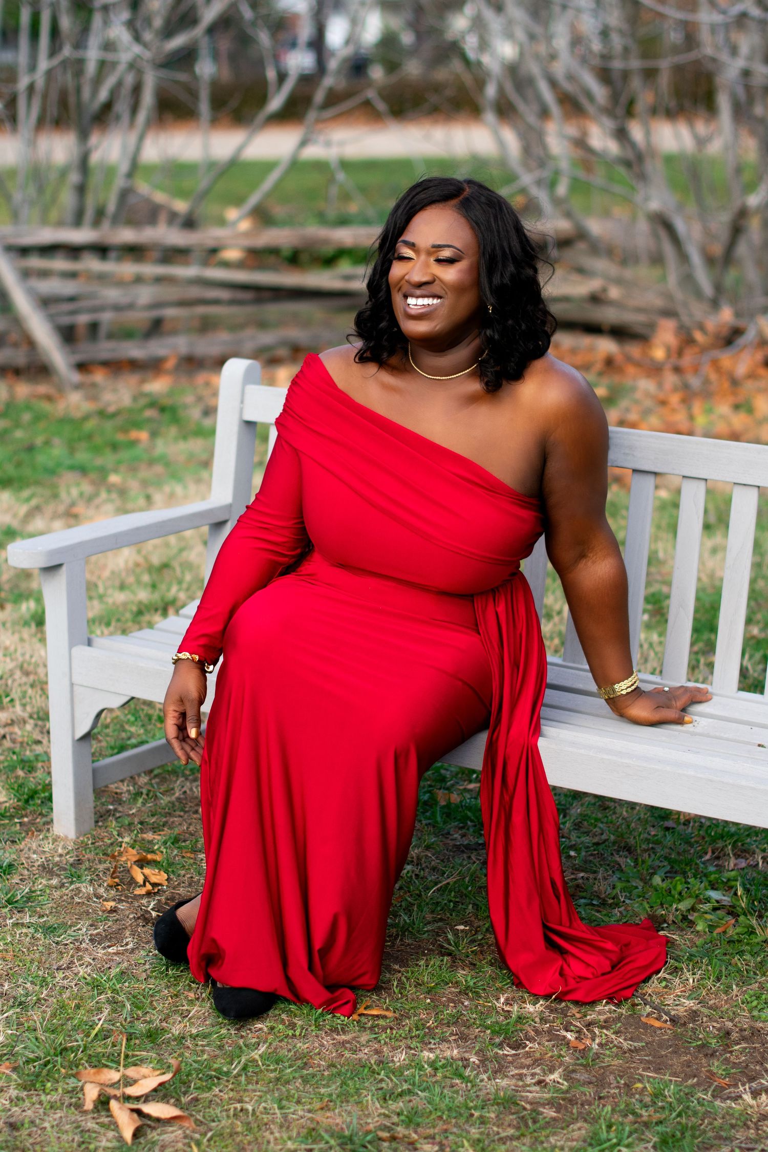 Woman wearing a glamorous one-shoulder red dress sits on a white wooden bench in an autumn garden setting. Professional headshot photography