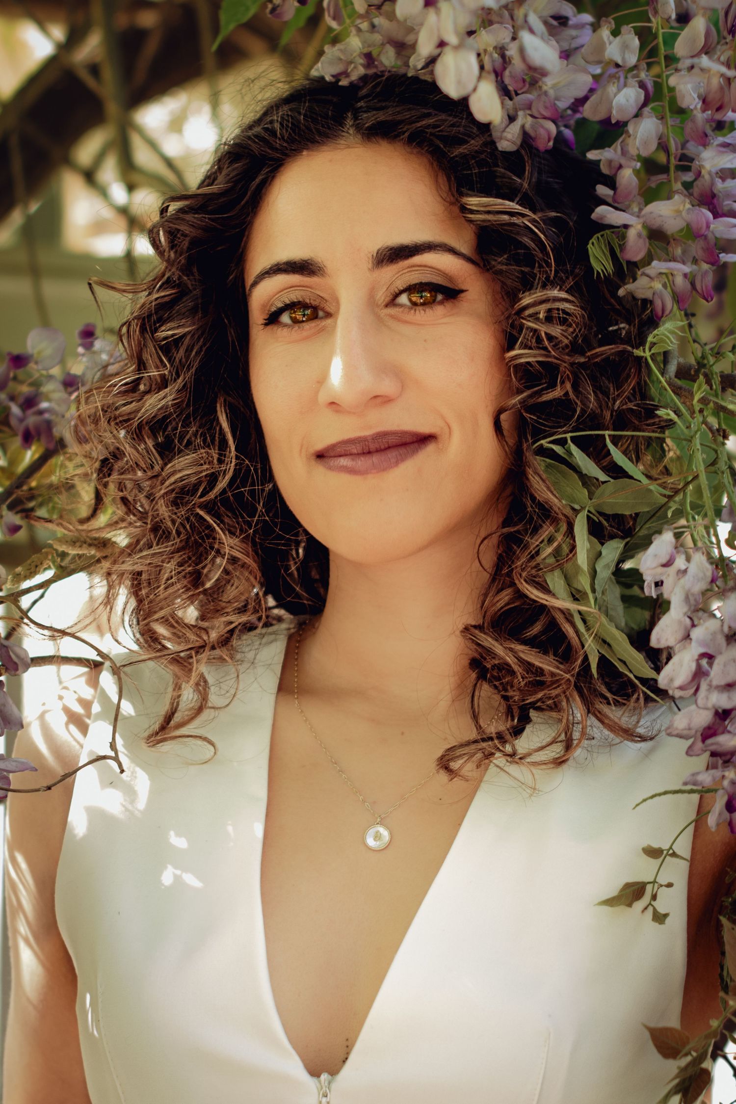 A woman in a white top with curly dark hair poses among blooming purple wisteria flowers. Professional portrait photography.