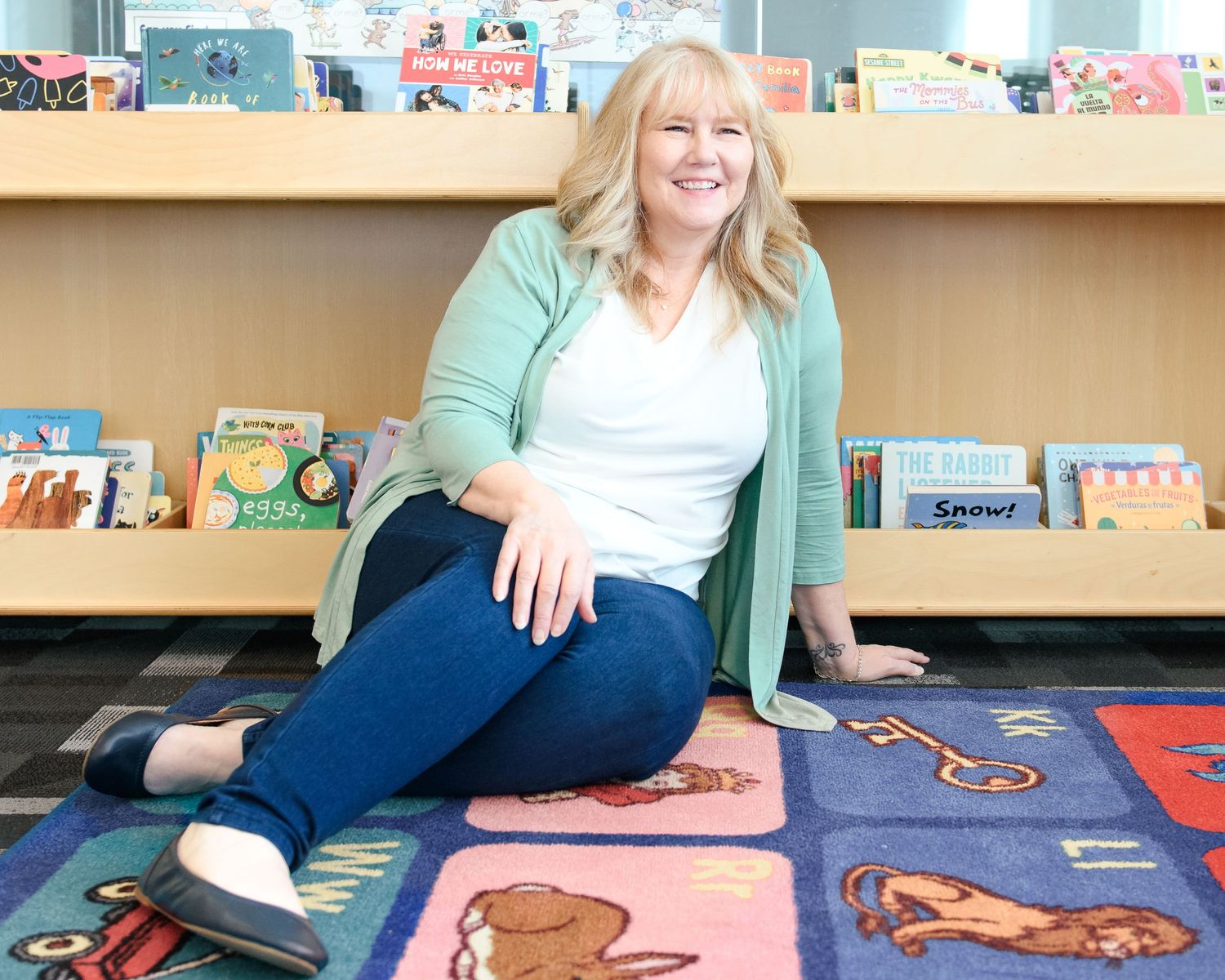 Blonde woman in green cardigan sitting on colorful rug in front of children's book display shelf.