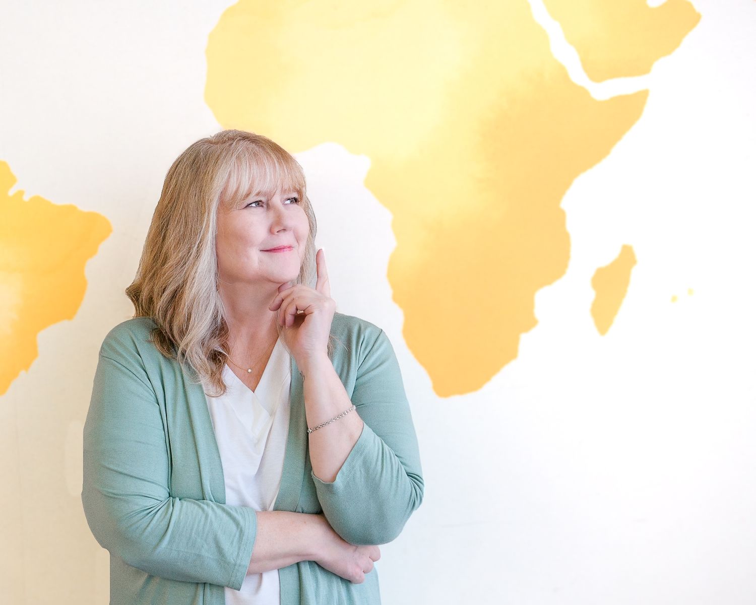 Smiling blonde woman in green cardigan standing thoughtfully before a large yellow Africa map mural.