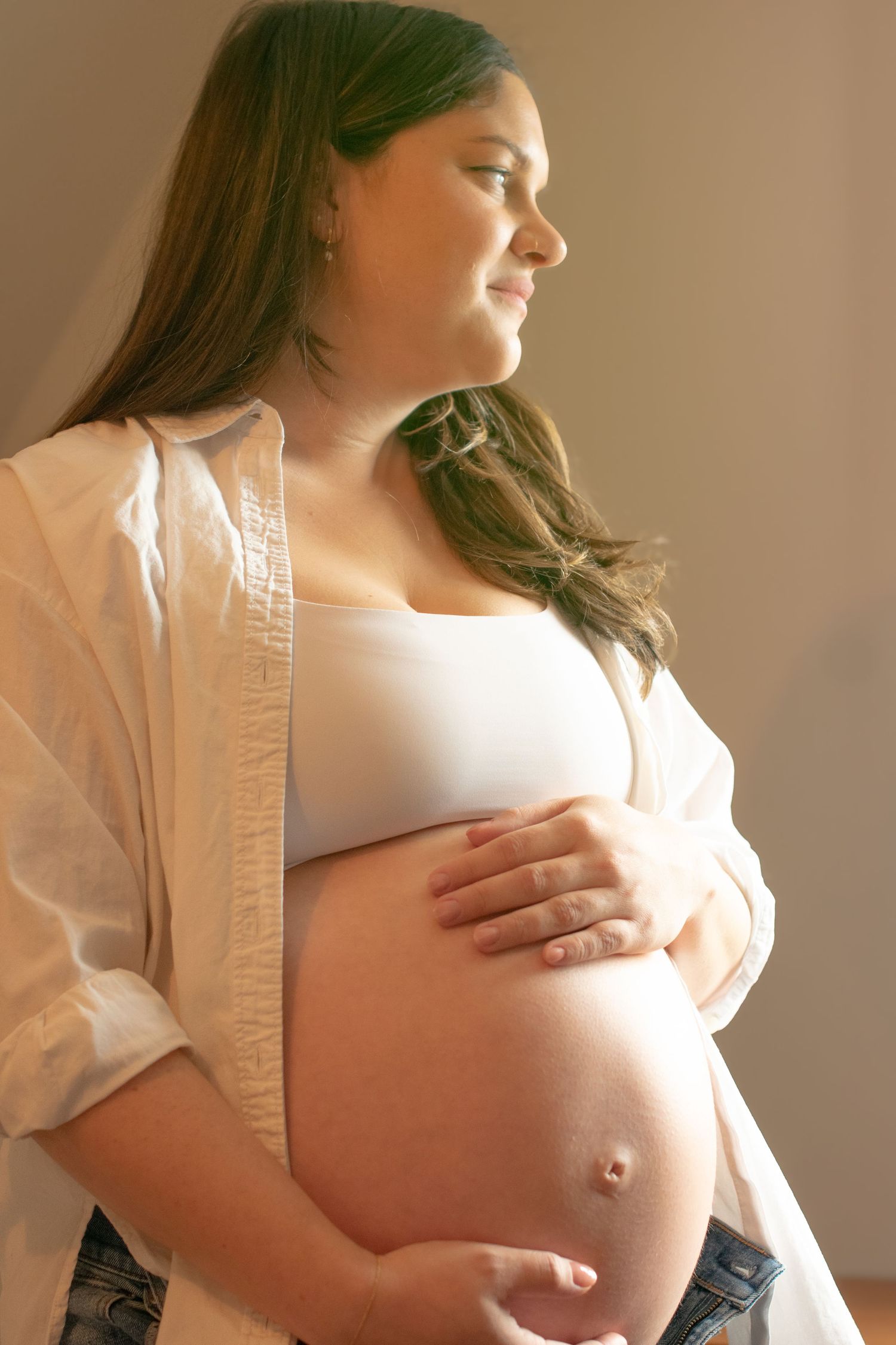 Side view maternity portrait with soft, warm light showing pregnant belly in casual white top and jeans.