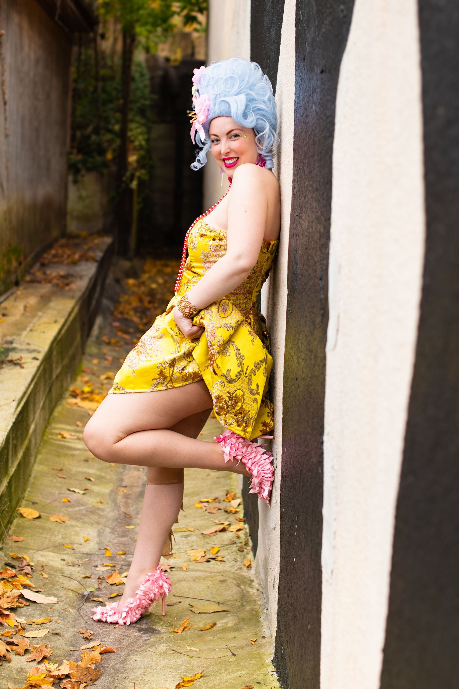 A vintage-inspired pin-up style photo with yellow baroque dress and pink bow heels posed against a black and white wall in an alley