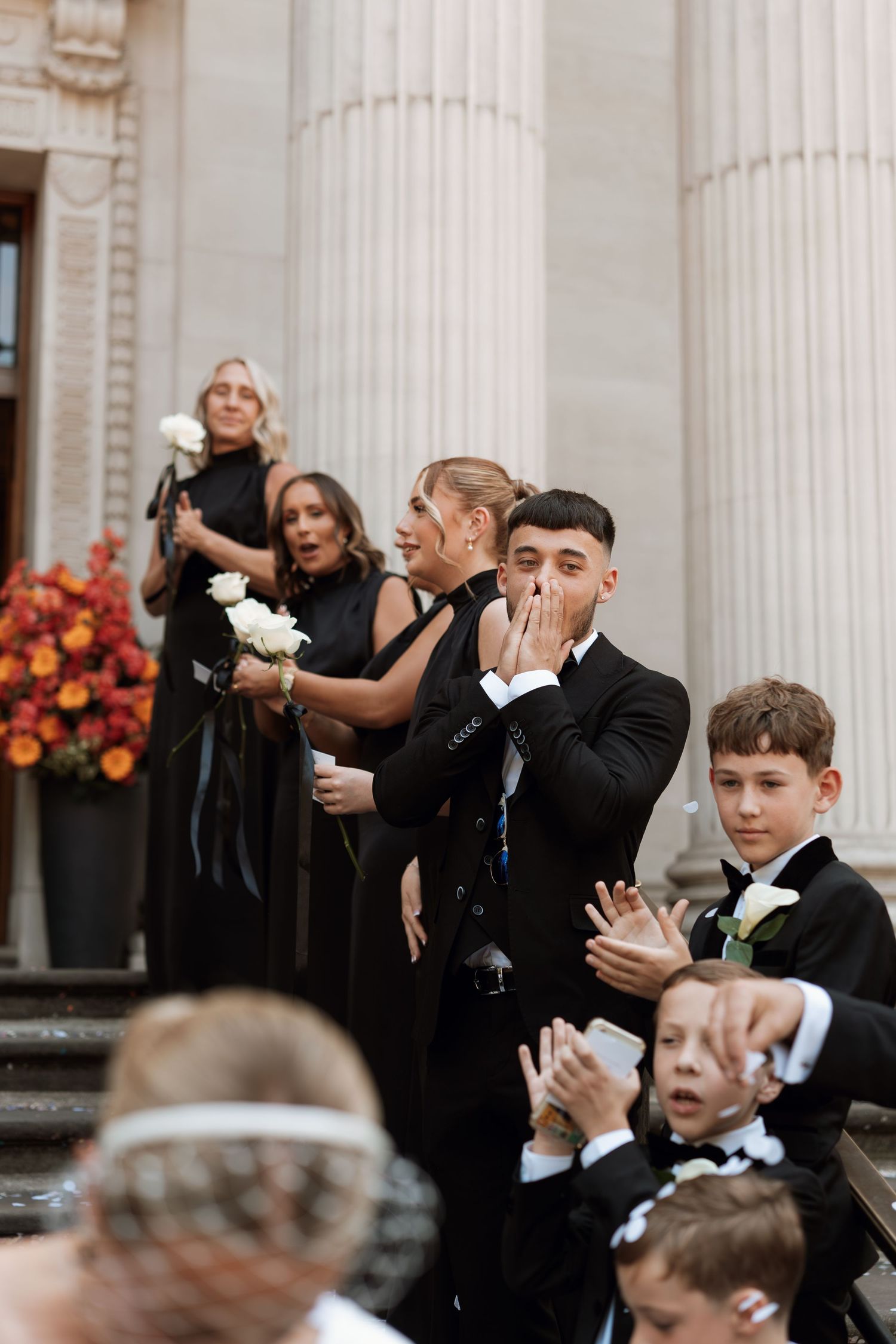 Wedding guests in formal attire clapping and celebrating during ceremony with white columns in background.