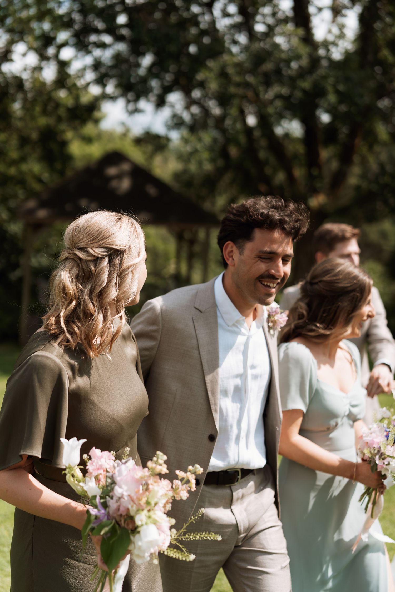 Wedding party members in sage green and beige attire walk together outdoors holding delicate floral bouquets.