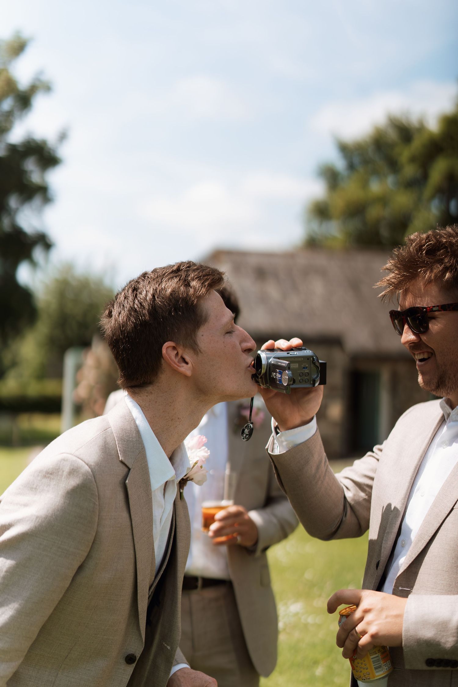 Two people in light colored suits sharing drinks and laughing at an outdoor event.