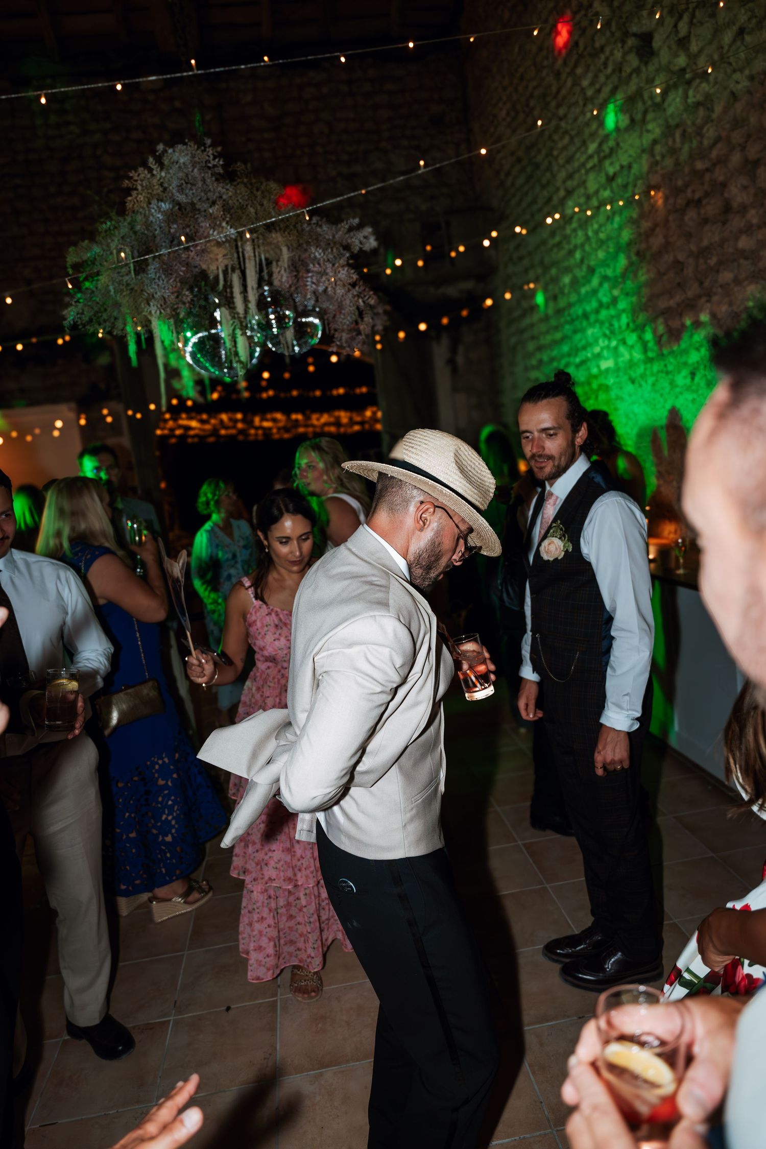 Wedding guests dance under colorful lights and string lighting at an indoor reception.