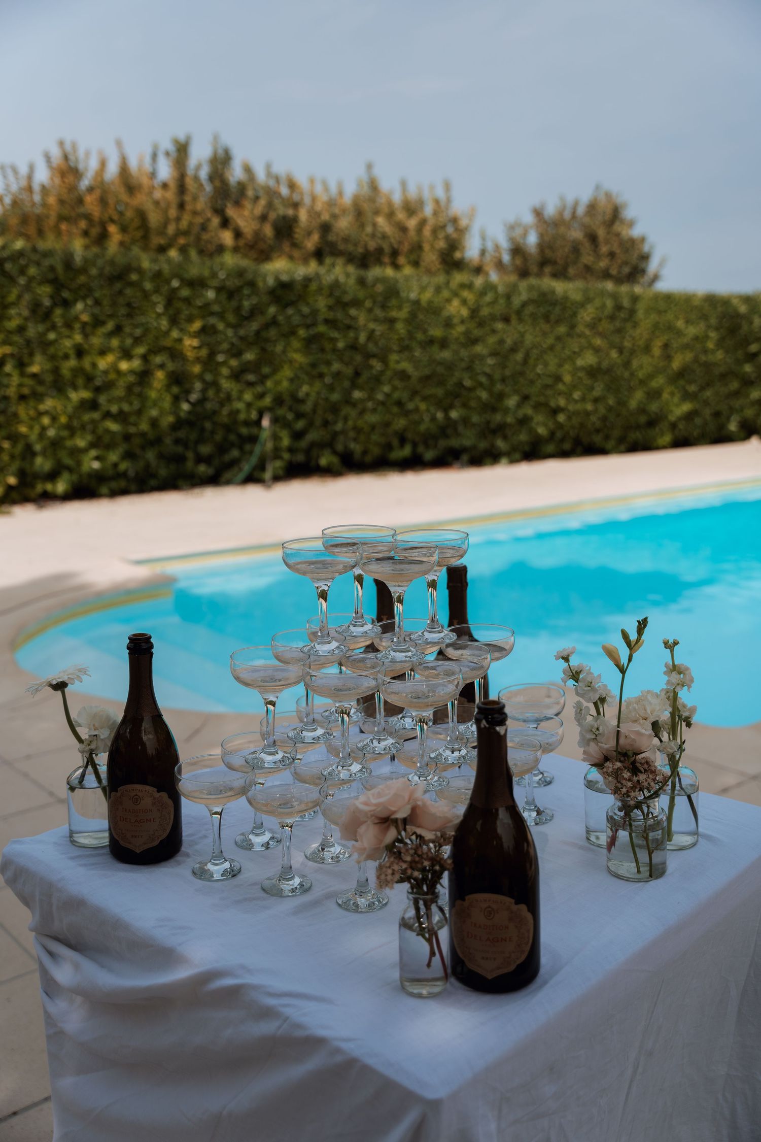 Champagne glasses and wine bottles arranged on a white tablecloth next to a swimming pool.