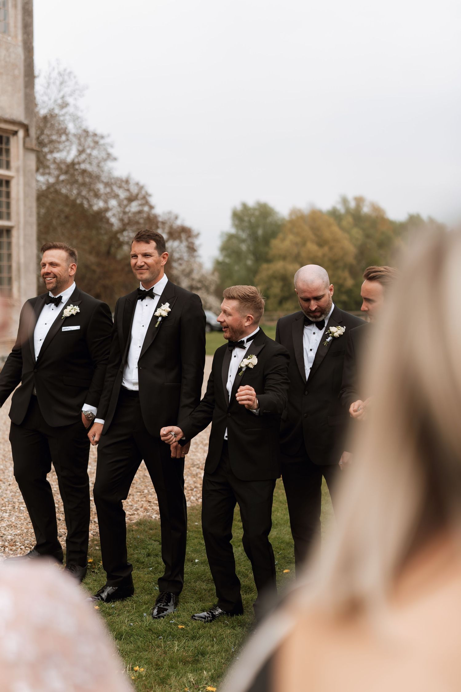 A group of groomsmen in black tuxedos share a candid laughing moment together on a grassy lawn.