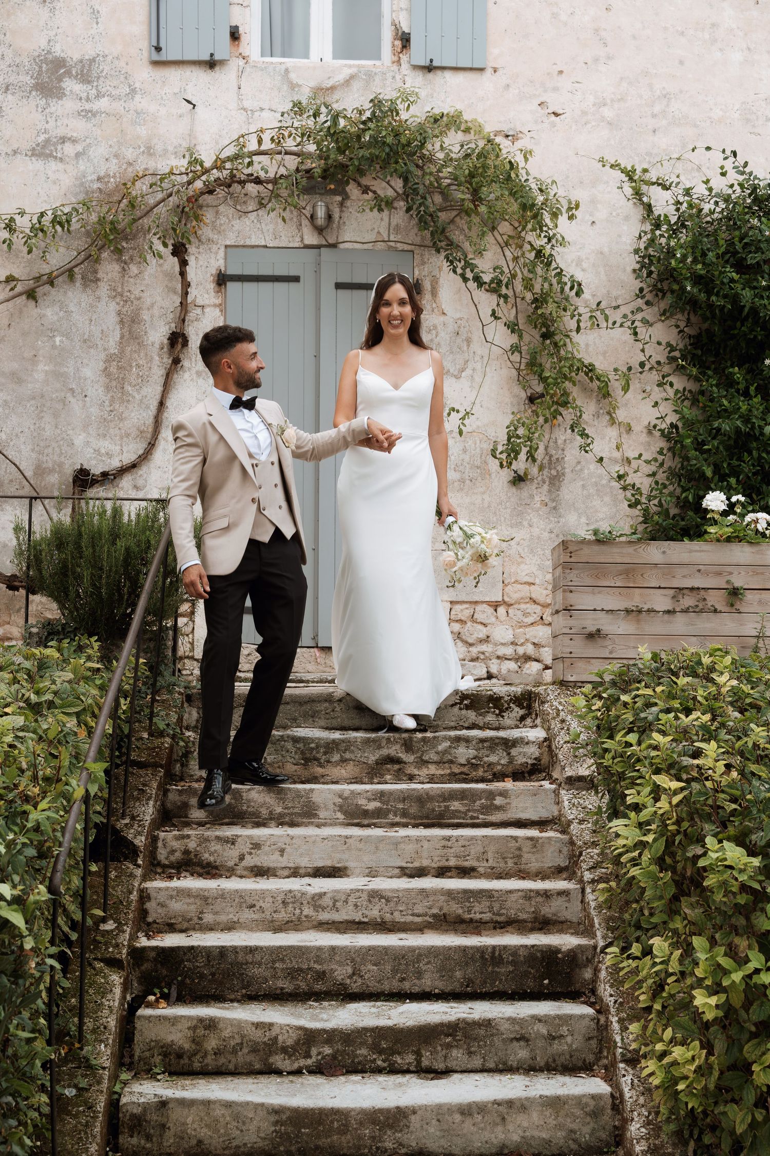 A couple poses on rustic stone steps against a weathered wall with climbing vines at a romantic outdoor venue.