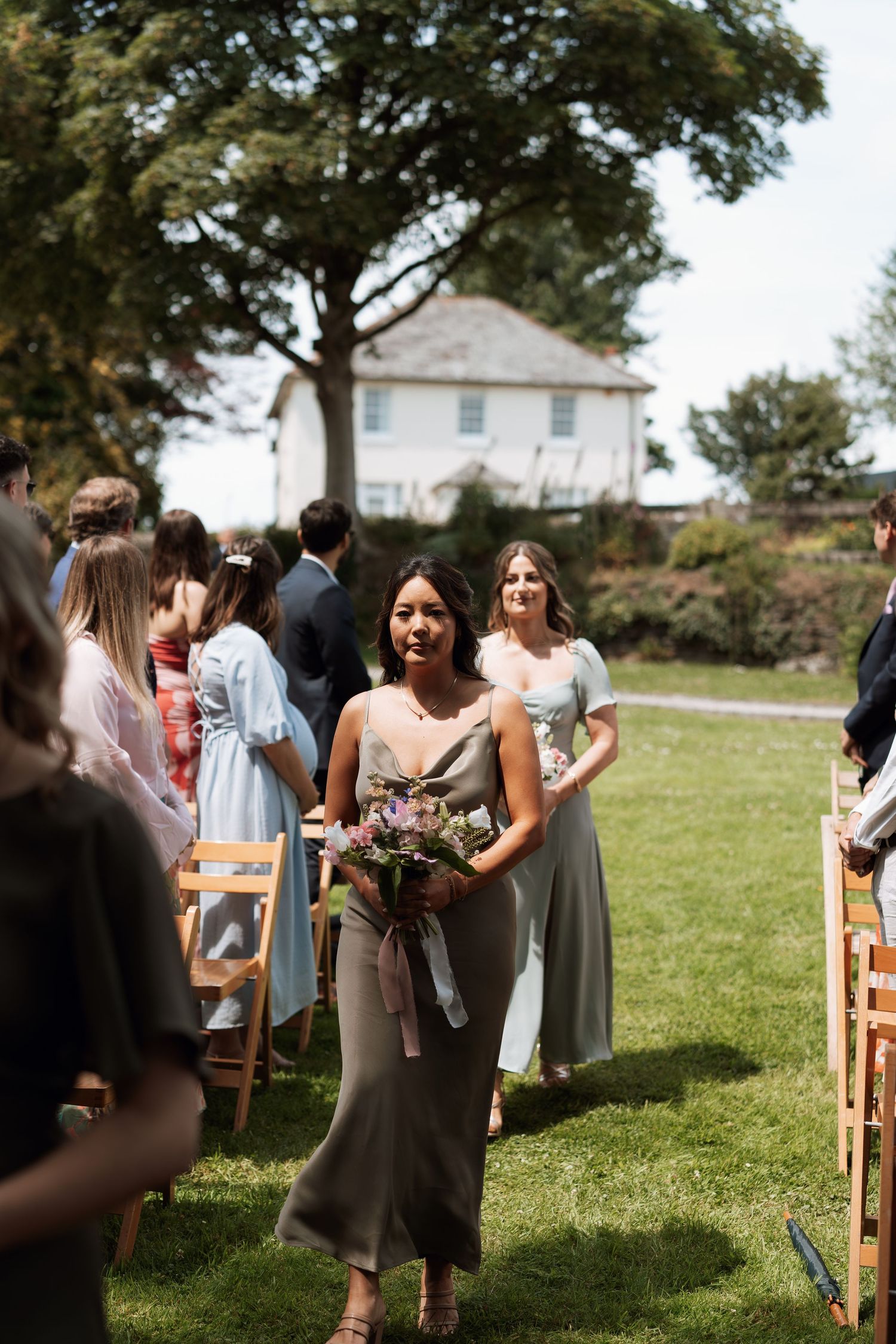 Wedding guests gathering outdoors on a sunny day with wooden chairs and green lawn.
