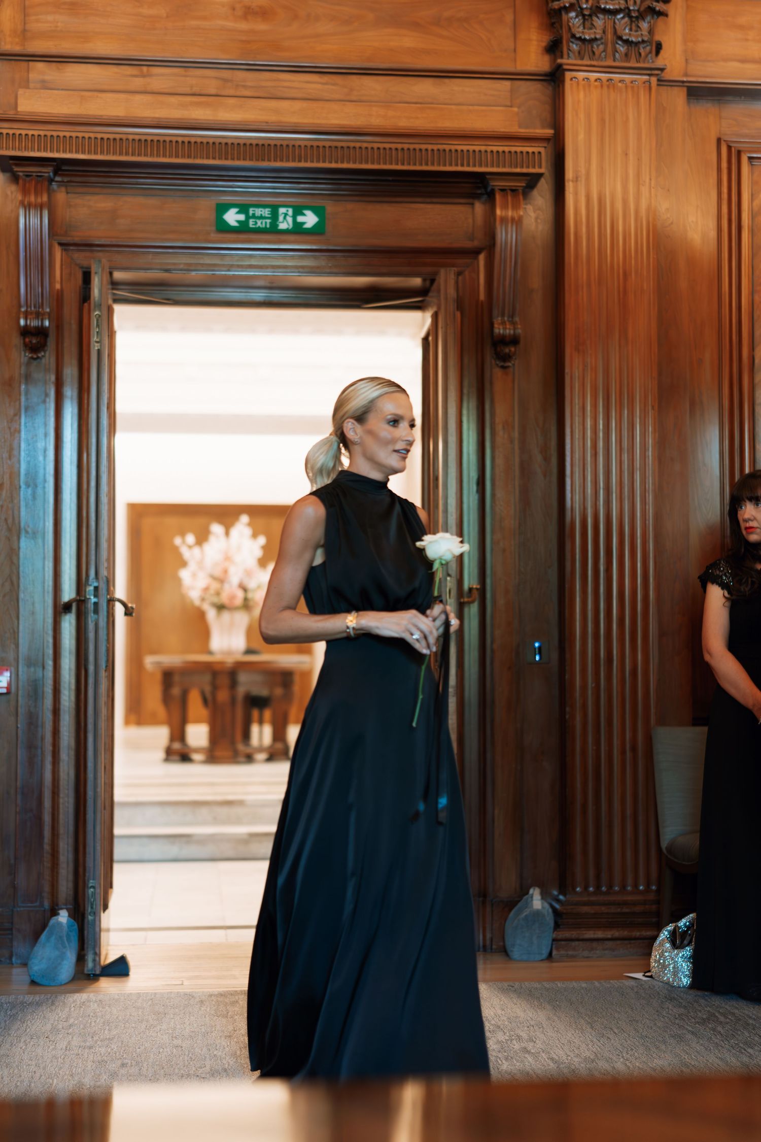 Elegant figure in black gown standing in ornate wooden doorway with exit sign above.