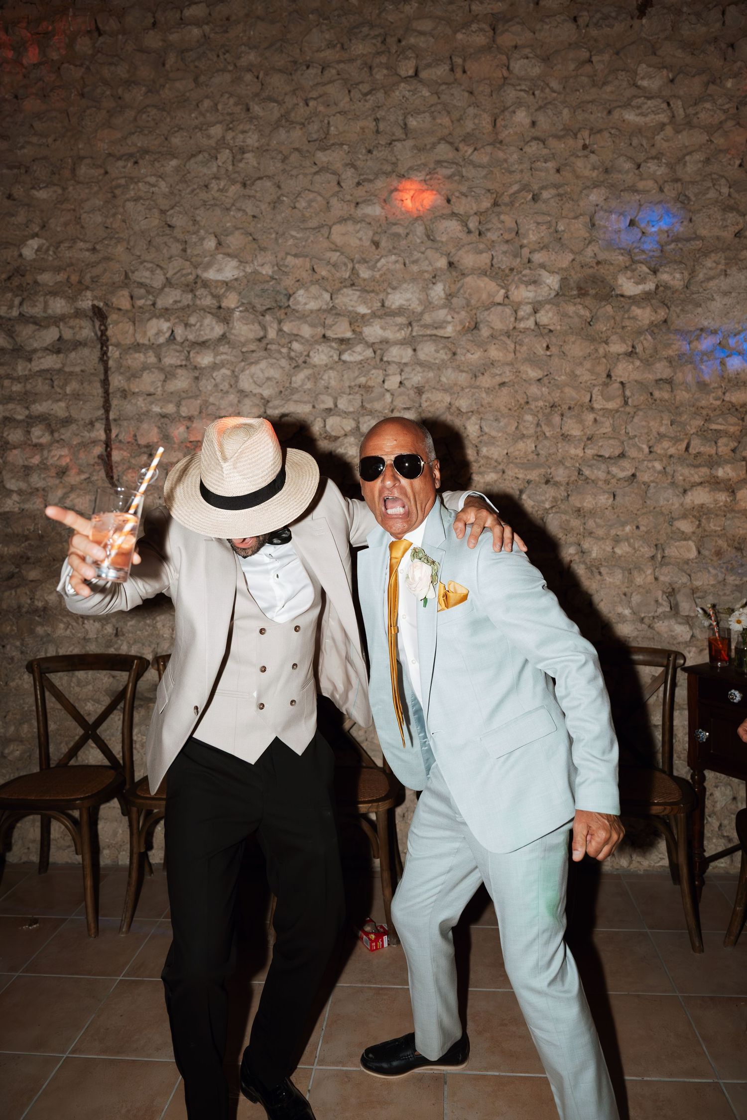 Party guests in formal wear dancing and posing enthusiastically against a stone wall.