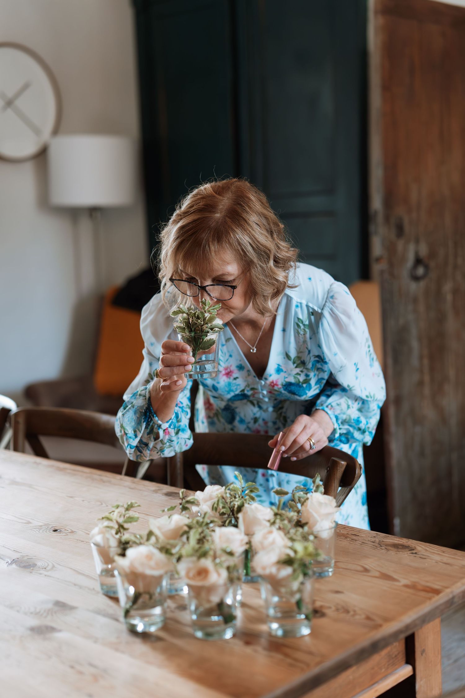 Person in floral blue top arranging delicate flower arrangements in small glass vases on wooden table.