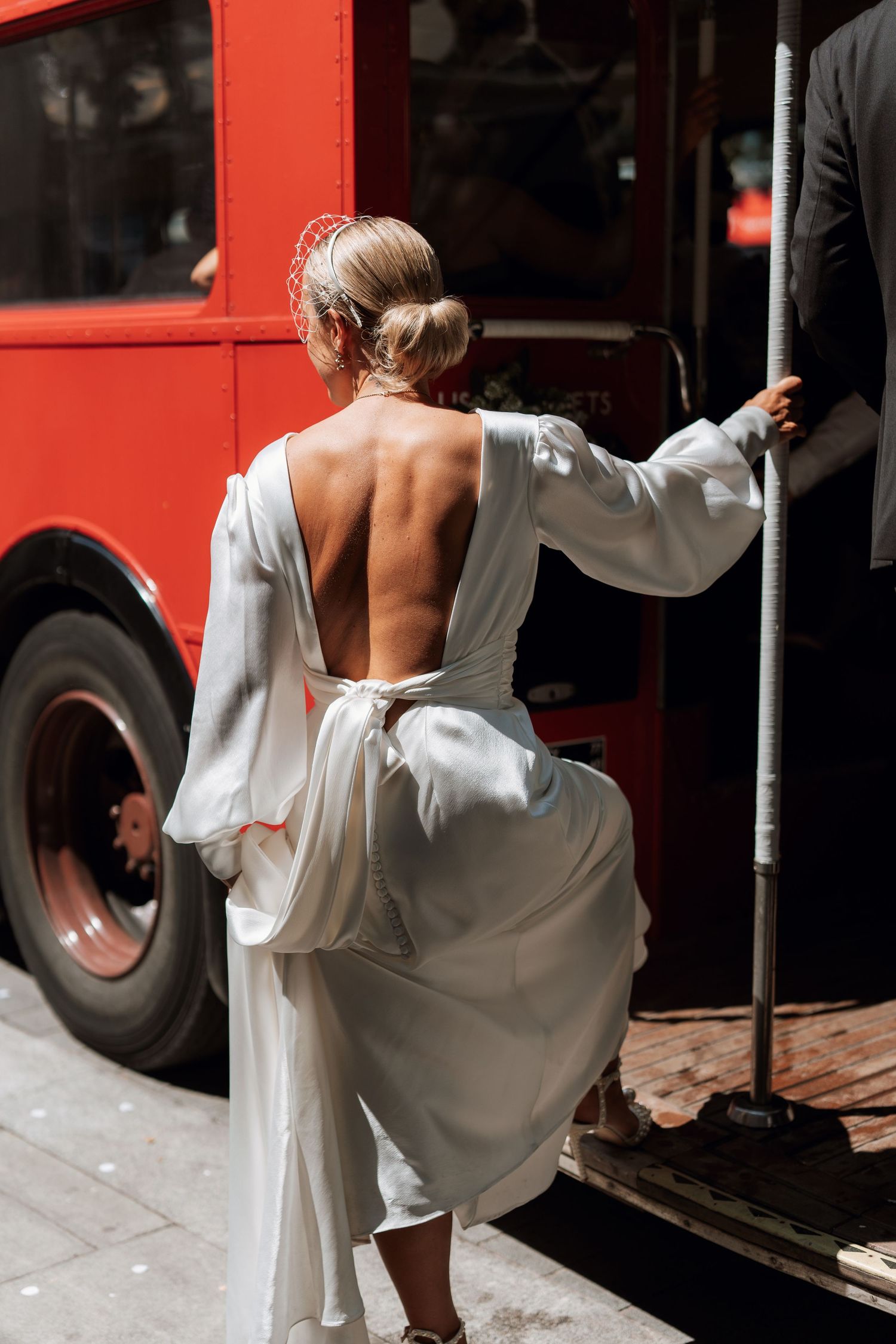 Woman in flowing white backless dress boards a red bus, showing elegant movement and style.