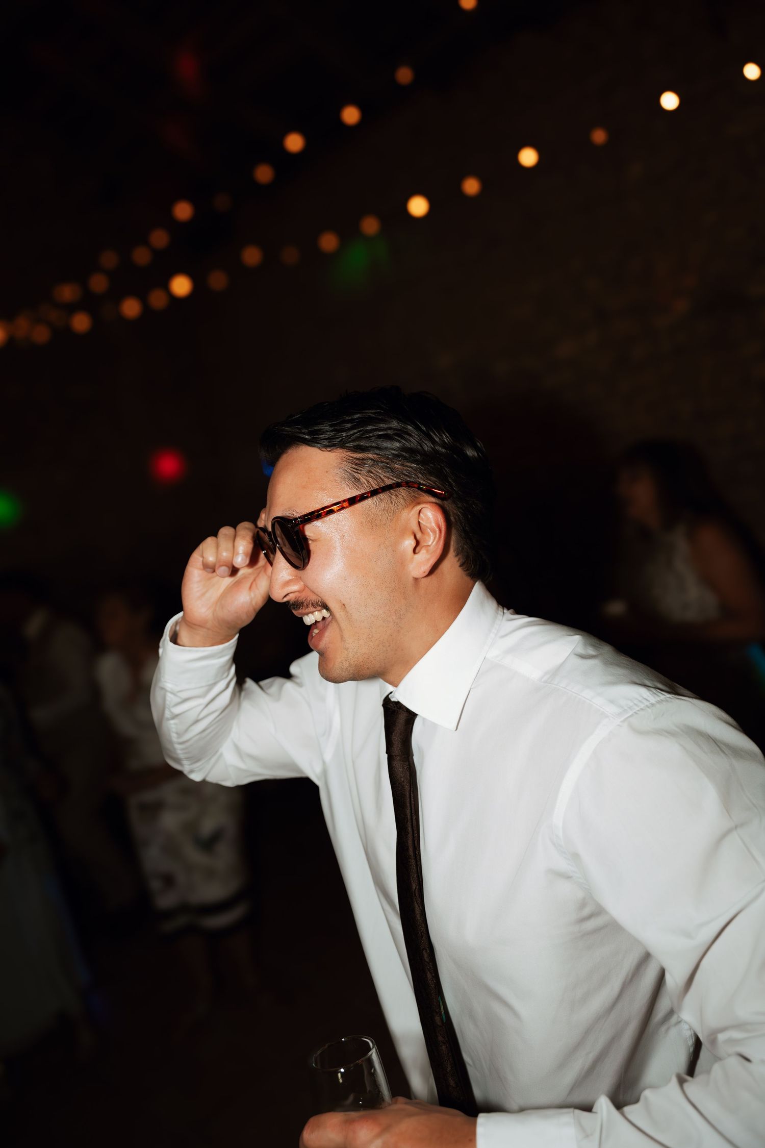 Person in white shirt and sunglasses dancing energetically at a party under colored lights.