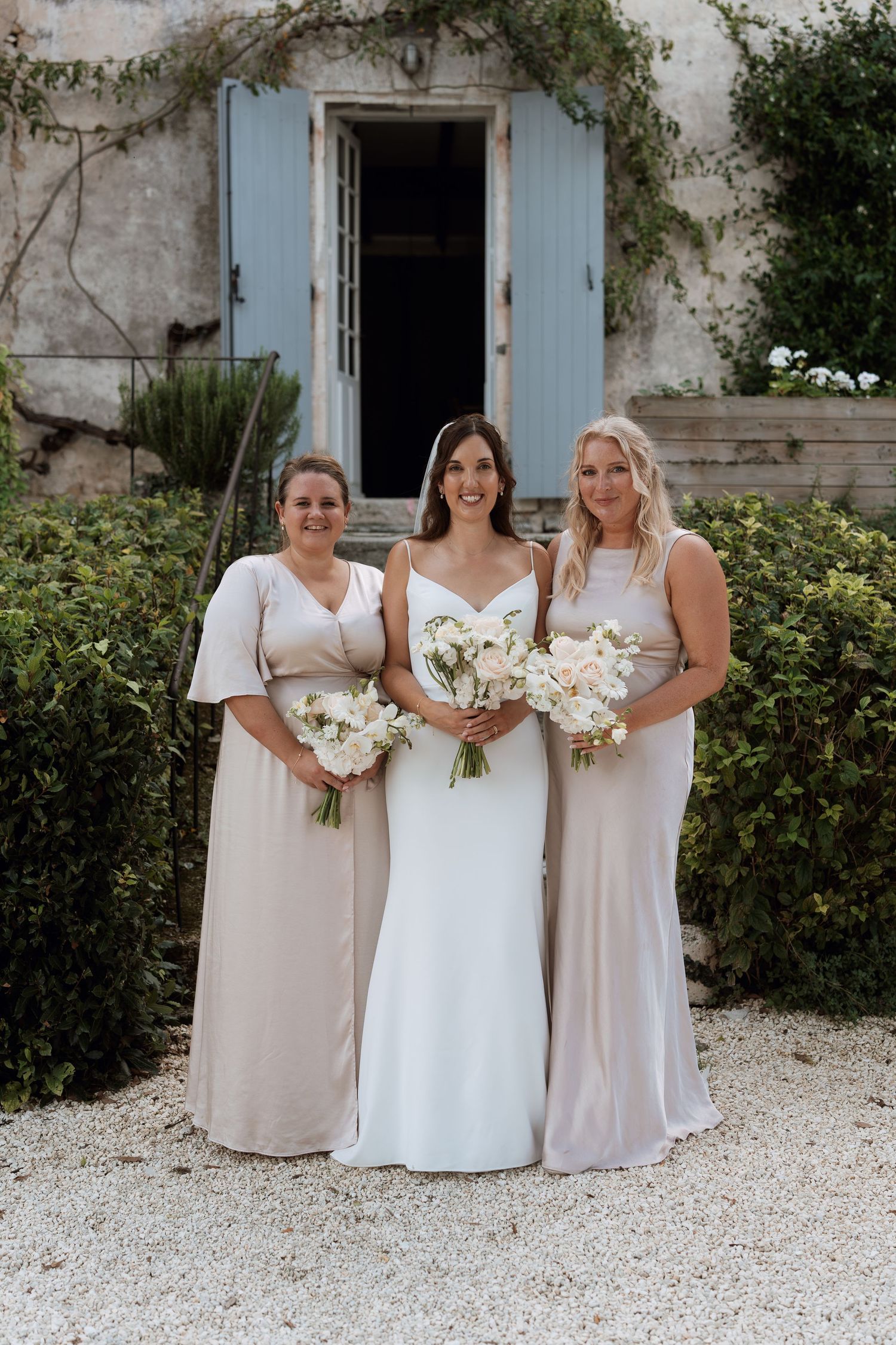 Bridal party in white and beige dresses pose with bouquets against stone building.