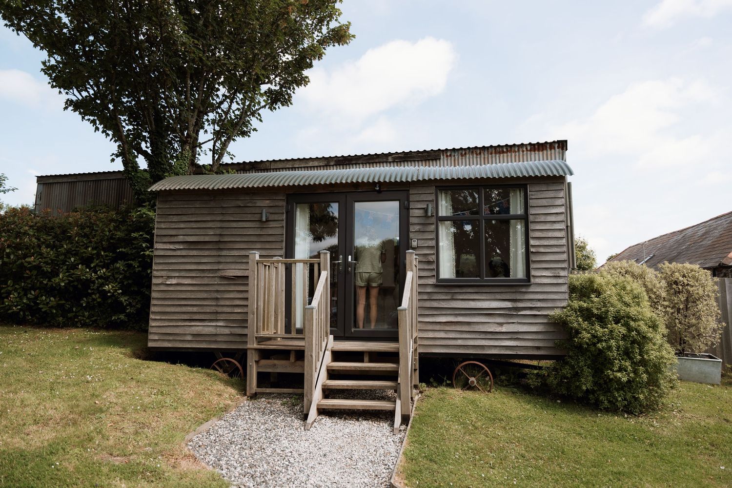 A modern wooden cabin with a gravel path and outdoor deck sits nestled among trees in a rural setting.