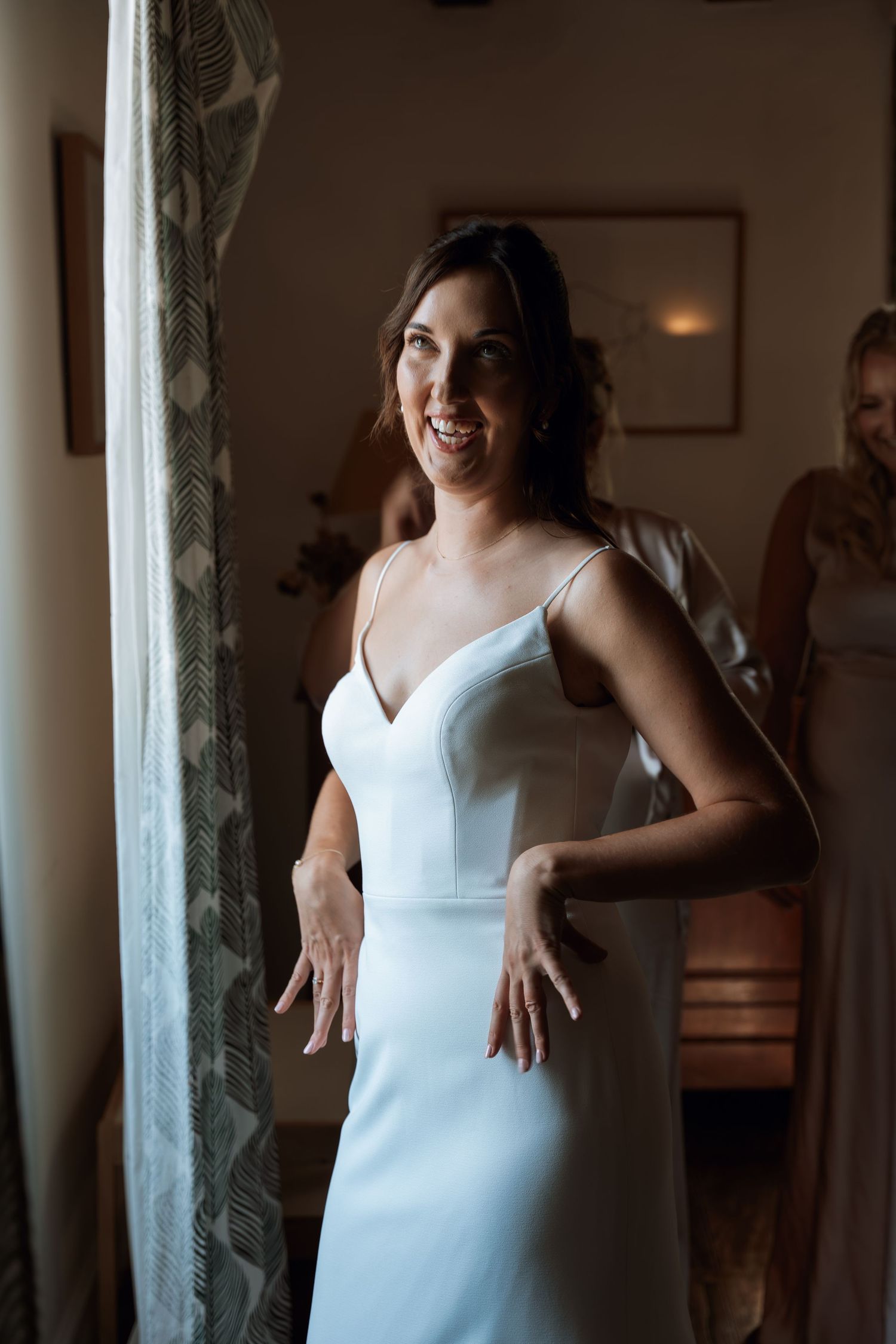 Woman in elegant white wedding dress poses by window in natural light.