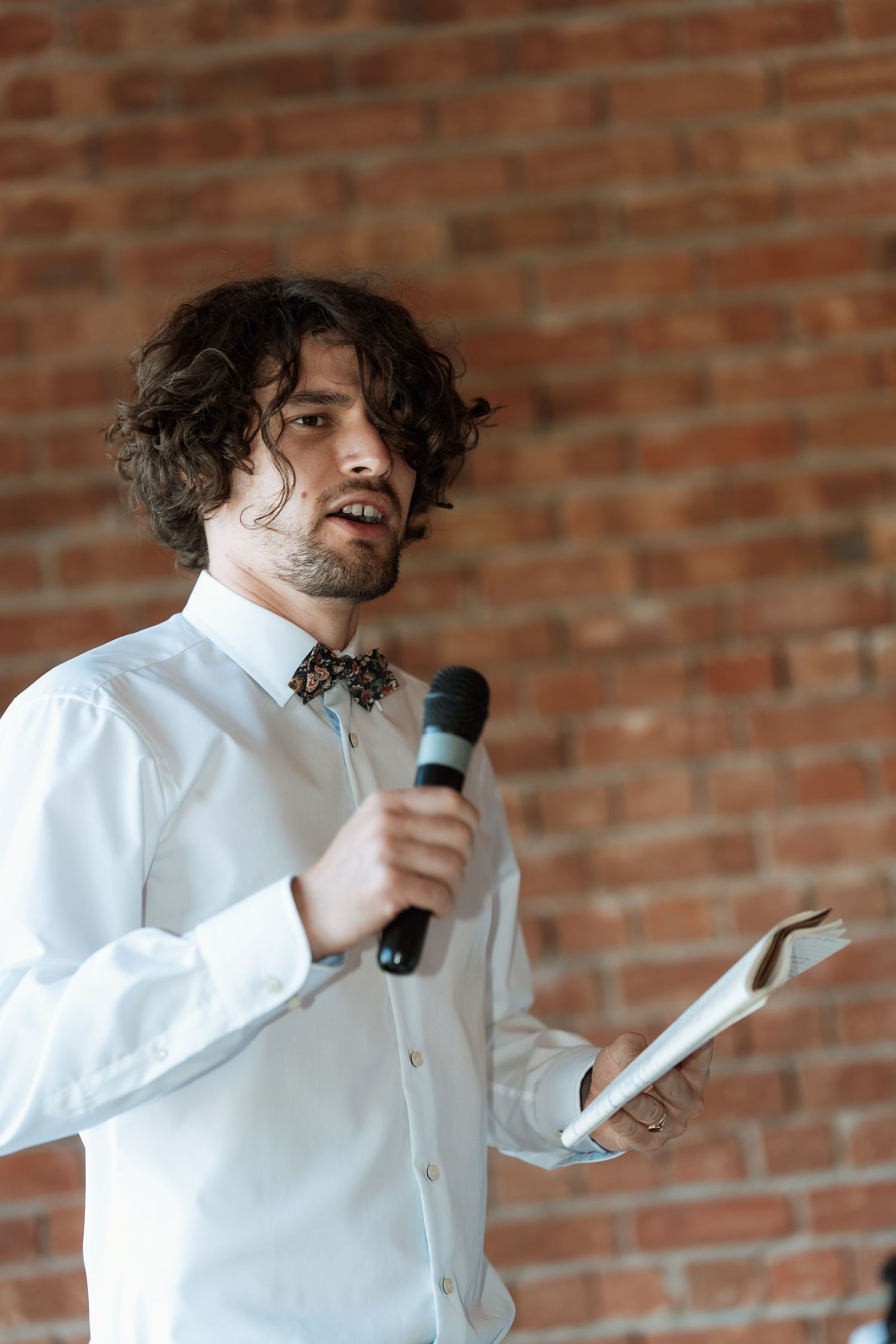 A speaker in a white shirt and bow tie delivers a speech against a brick wall backdrop.