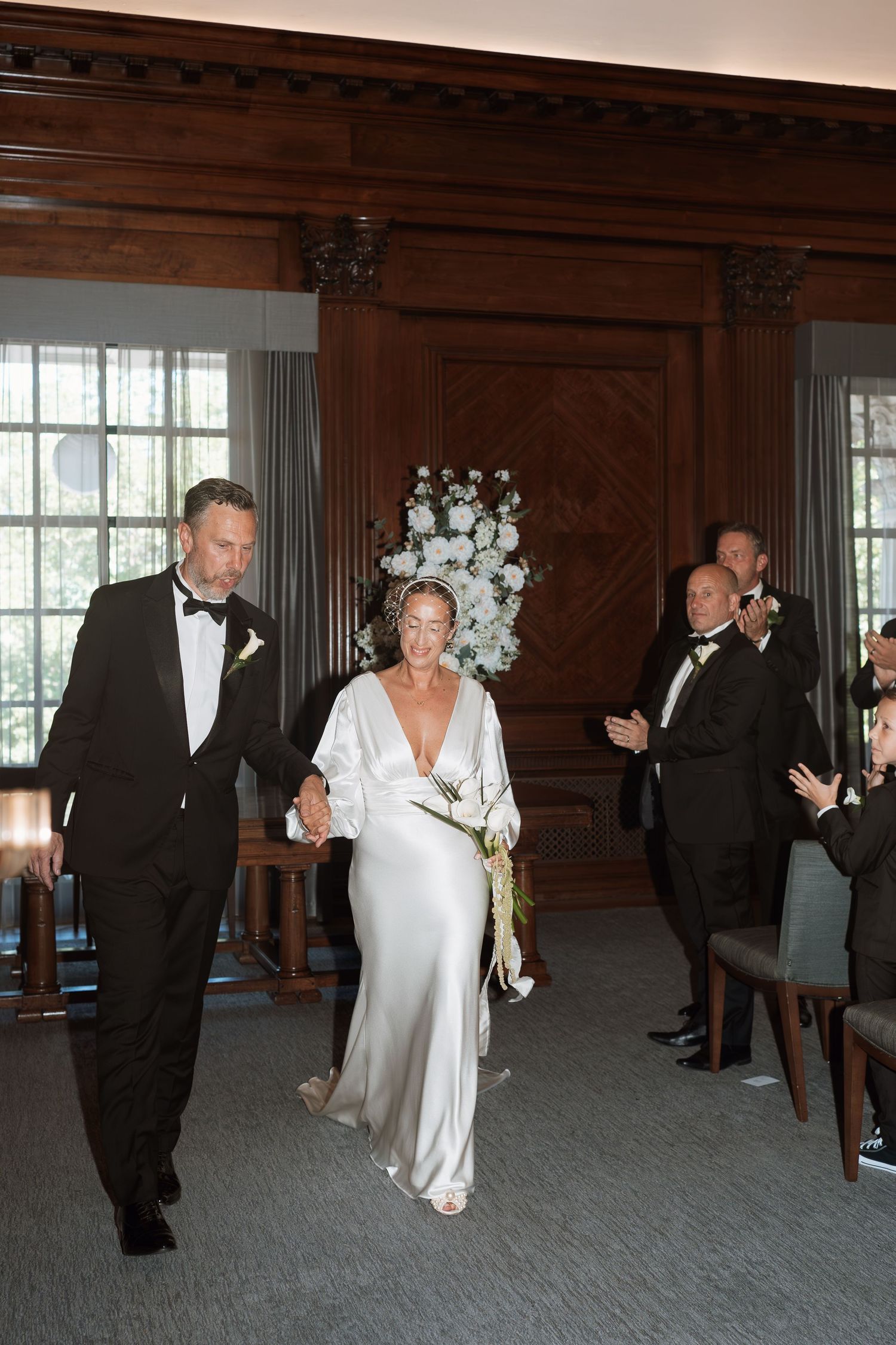Wedding party members in formal attire dancing in a grand wood-paneled ballroom.