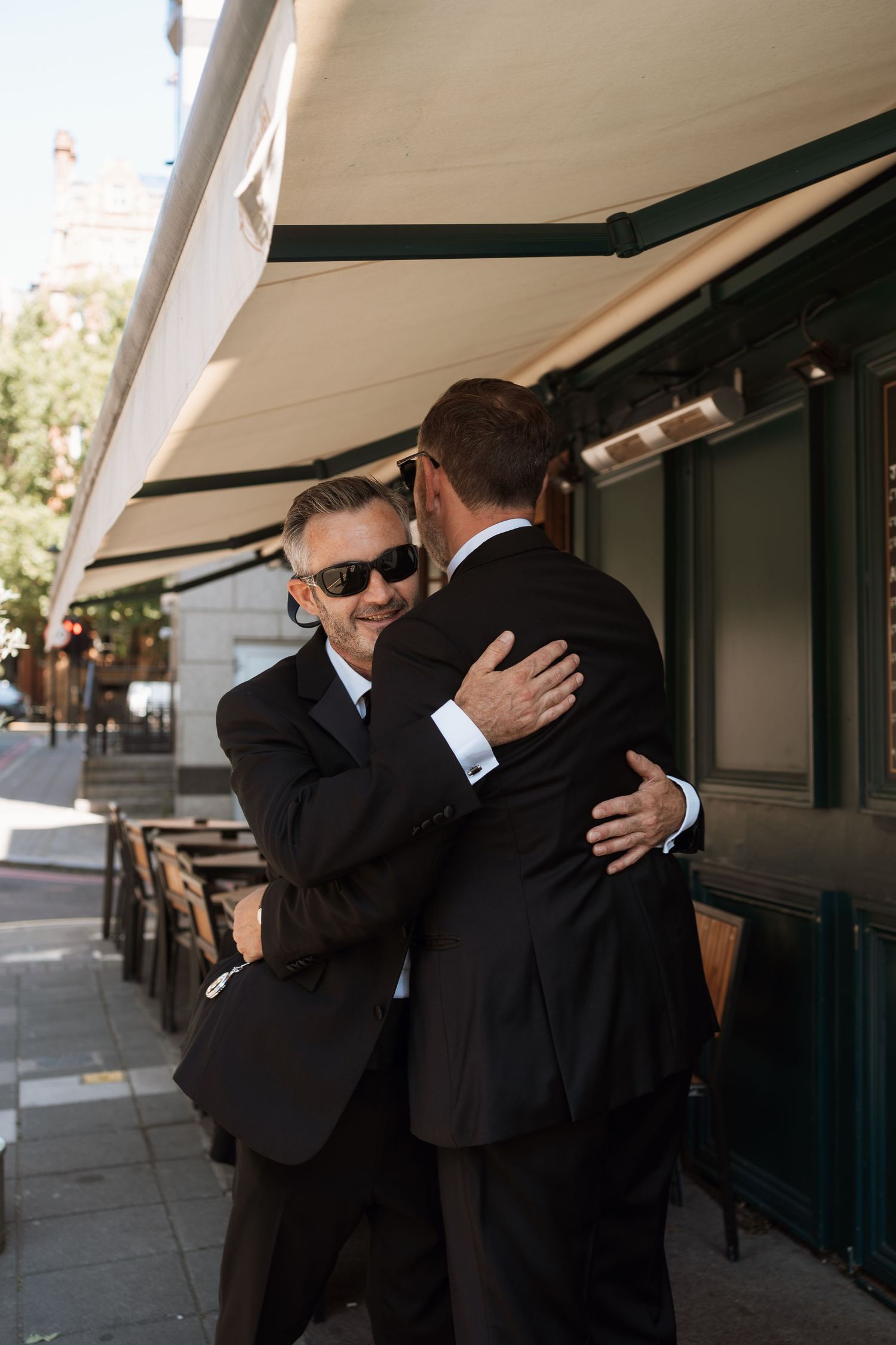 Two men in black suits and sunglasses embrace emotionally in a series of connected photos outside a building.