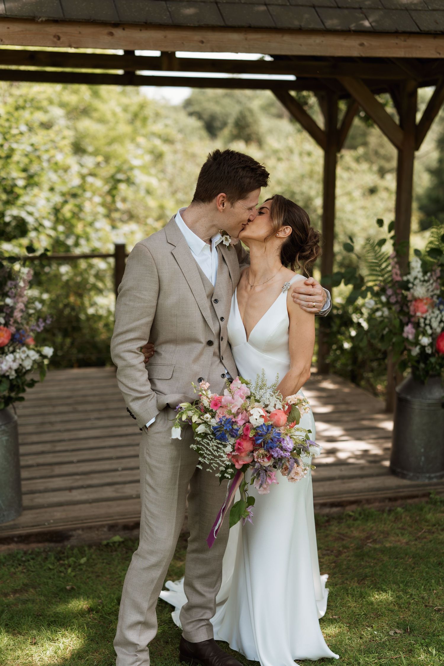 Romantic wedding couple shares a kiss under a rustic wooden pavilion with floral bouquet.